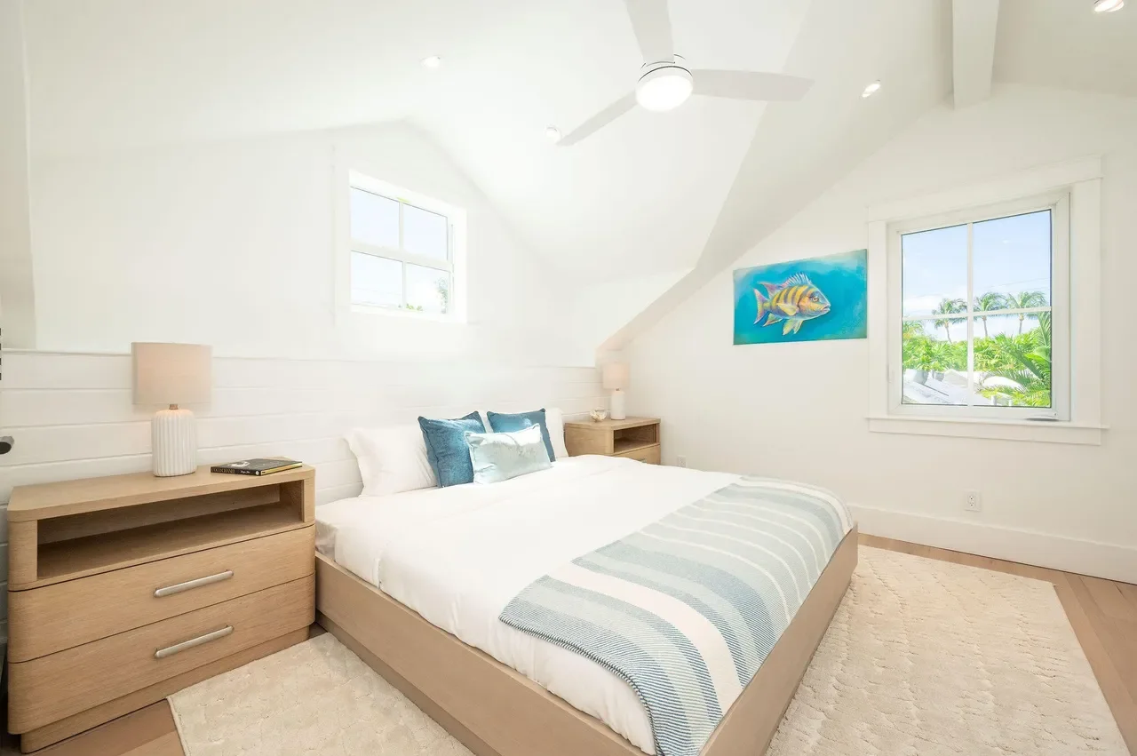 Light-filled Key West bedroom with coastal artwork, natural wood bed, white walls, ceiling fan, and windows framing lush tropical greenery.