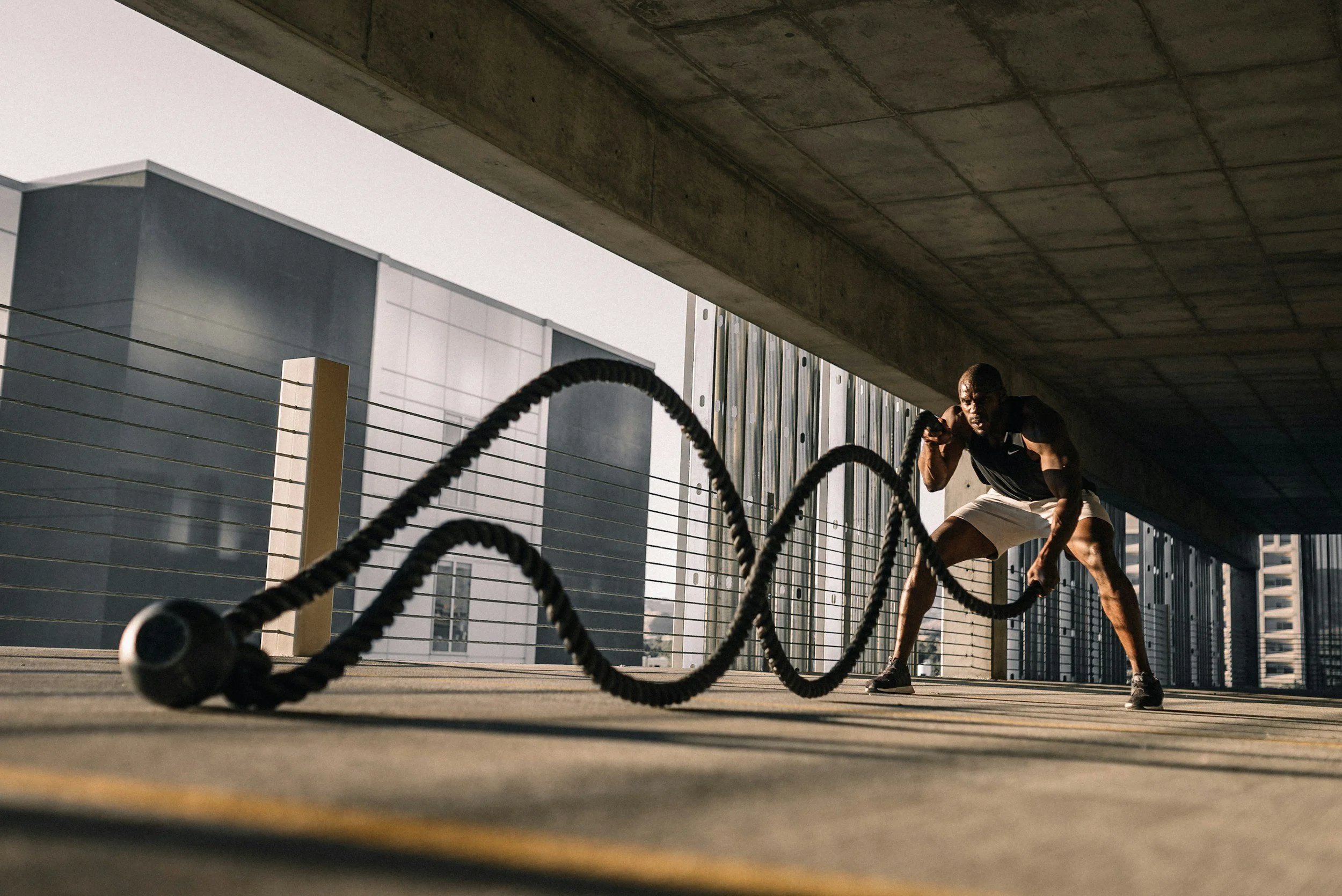 Homem de cabelo curto faz exercício de crossfit com corda de batalha em ambiente ao ar livre, com prédios ao fundo.