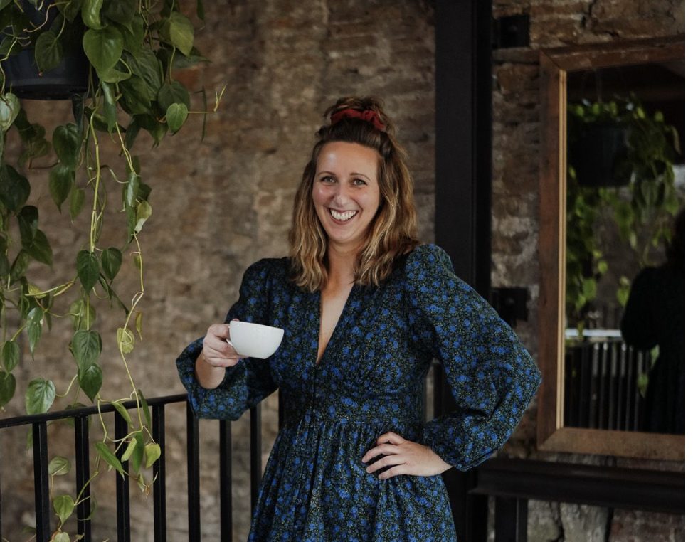 A woman with wavy light brown hair, wearing a blue floral dress, smiling and holding a white cup, standing indoors near a plant with hanging leaves and a mirror.
