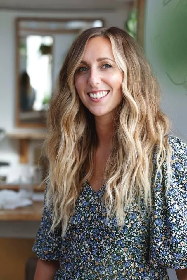 A smiling woman with long, wavy blonde hair wearing a floral dress, standing indoors with a mirror and greenery in the background.