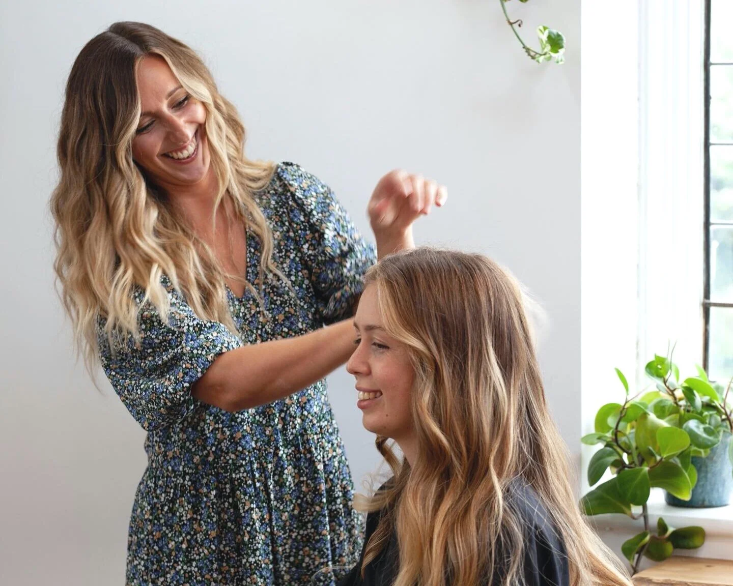 A woman with long, wavy blonde hair is laughing and happily styling a young woman's long, wavy hair in a bright room with a large window and green houseplants.