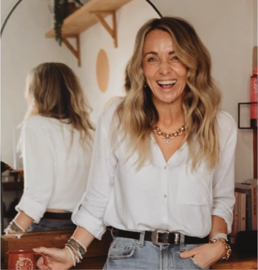 A woman with wavy blonde hair smiling and looking to the right, standing in a room with a mirror, wooden shelf, and books. She is wearing a white shirt, jeans, and gold jewelry.