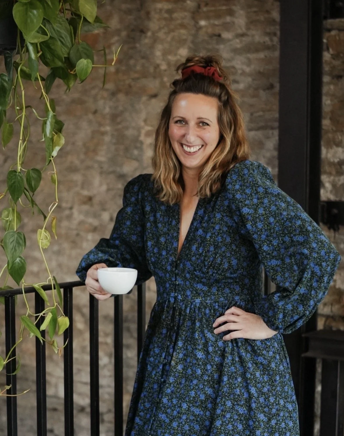 A woman with wavy, shoulder-length hair, held back with a red hair tie, smiling and holding a white coffee cup in her right hand. She is wearing a long-sleeved, dark blue dress with a floral pattern. She is standing next to a black metal railing, with a brick wall in the background, and a hanging green plant to her left.