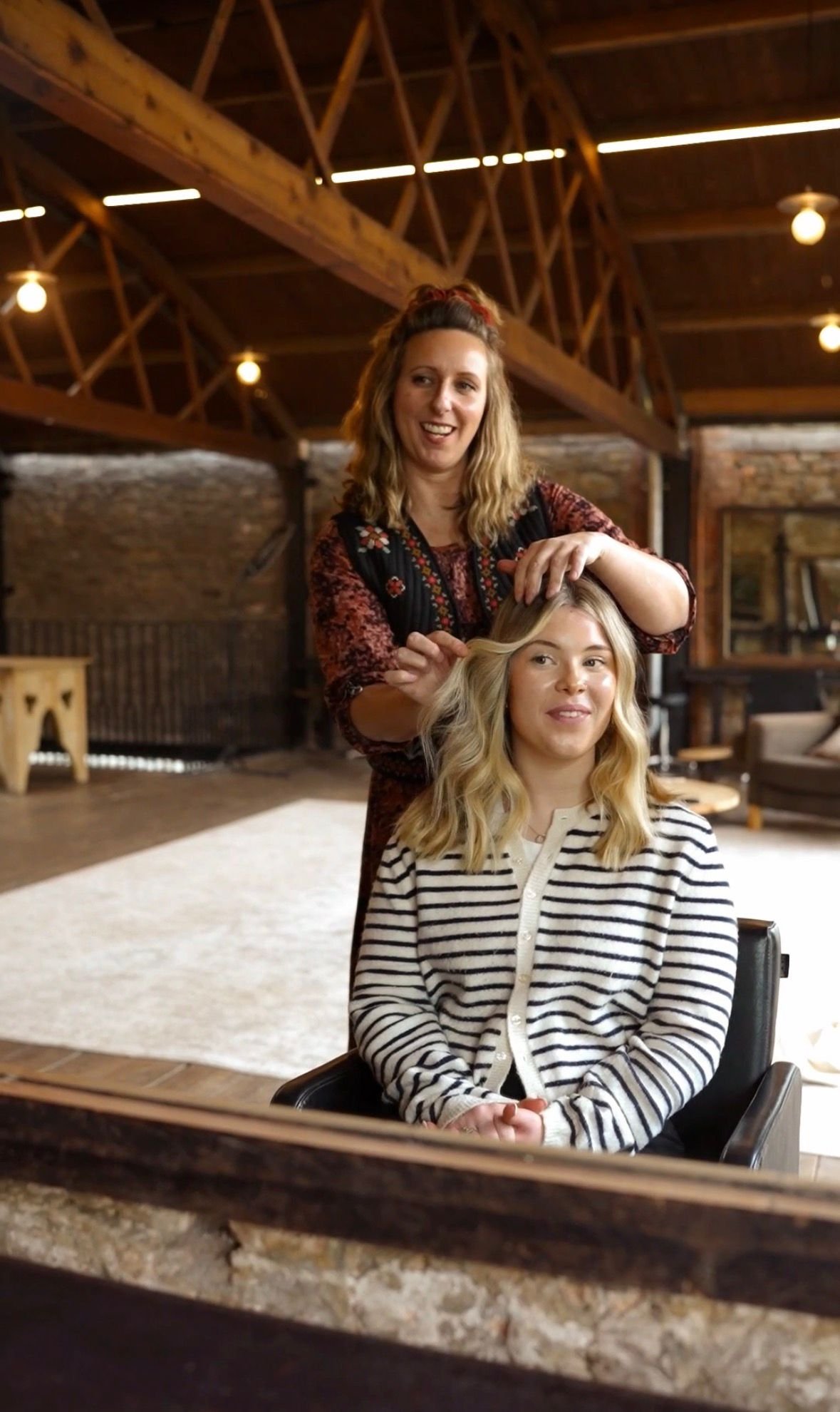 A woman is sitting in a salon chair, getting her hair styled by a stylist in a rustic, warmly lit salon with wooden beams and brick walls.