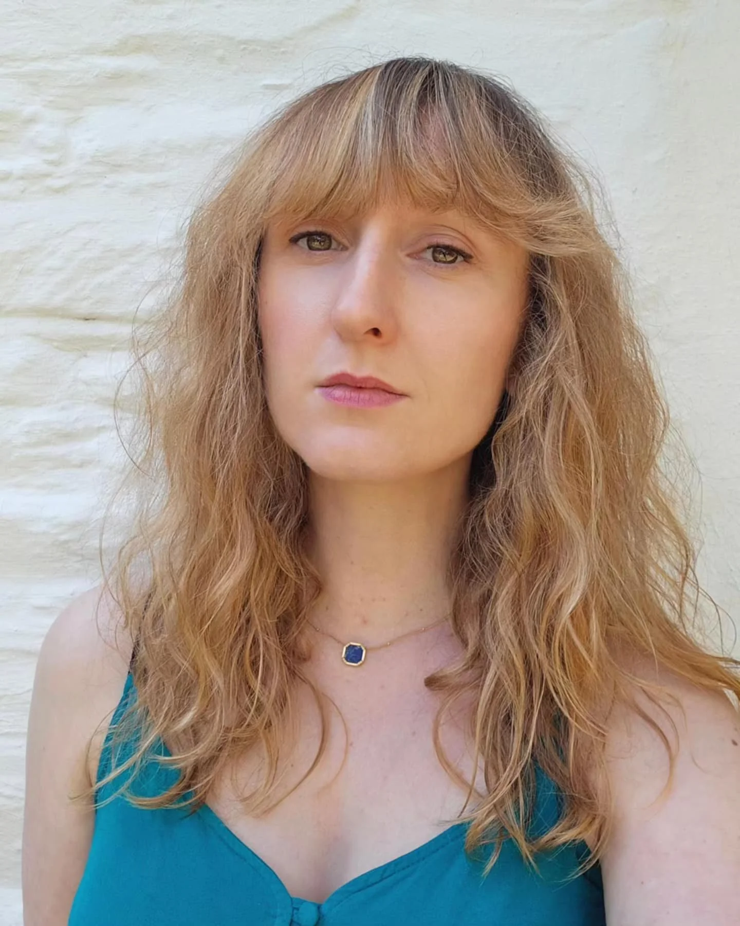A woman with wavy, red hair and fair skin wearing a blue top and a gold necklace with a blue gemstone, standing outdoors against a white brick wall.