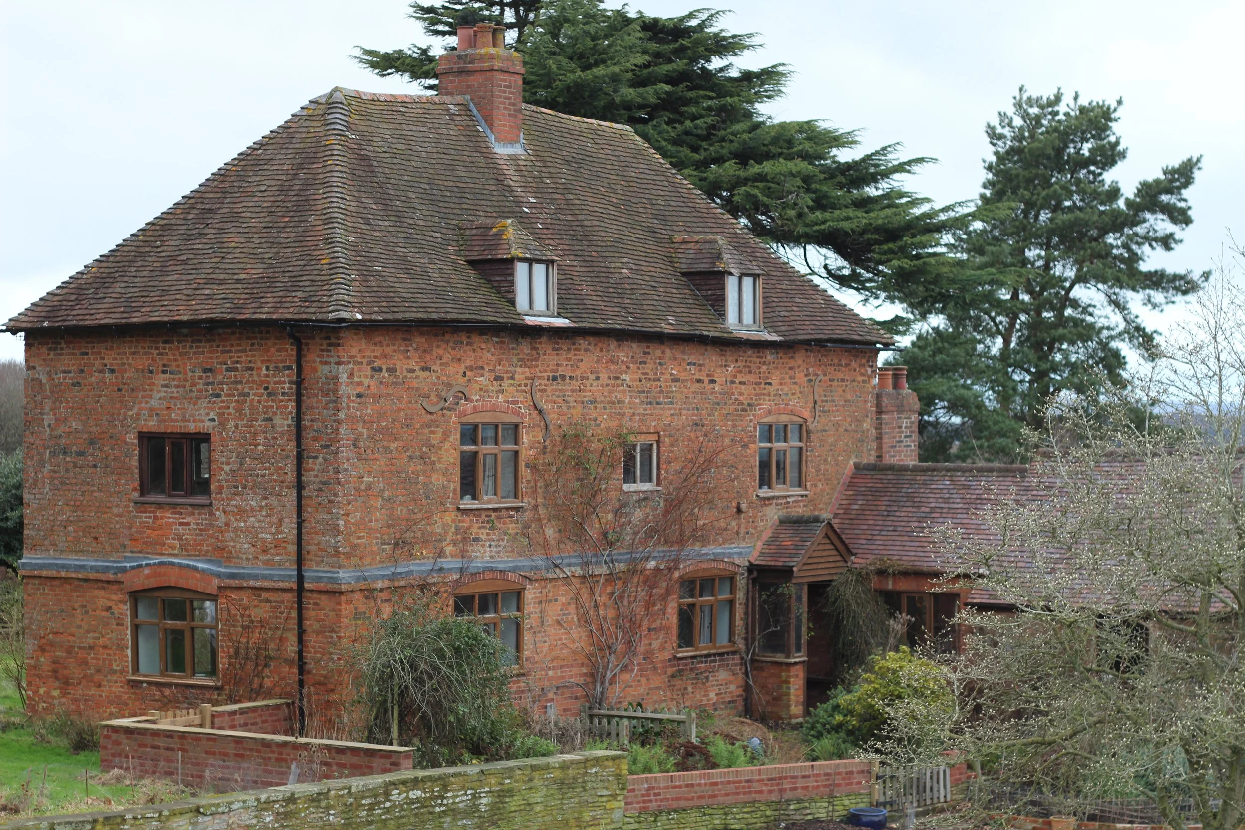 A large brick house with a pitched roof, multiple small windows, and surrounding trees and shrubs.