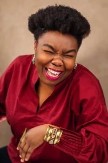 Woman with short curly hair laughing, wearing a red satin blouse and gold jewelry.