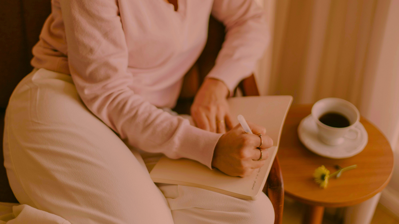 A person sitting on a chair writing in a notebook with a pen, next to a small round wooden table with a cup of coffee and a yellow flower. Represents a healthy morning routine.