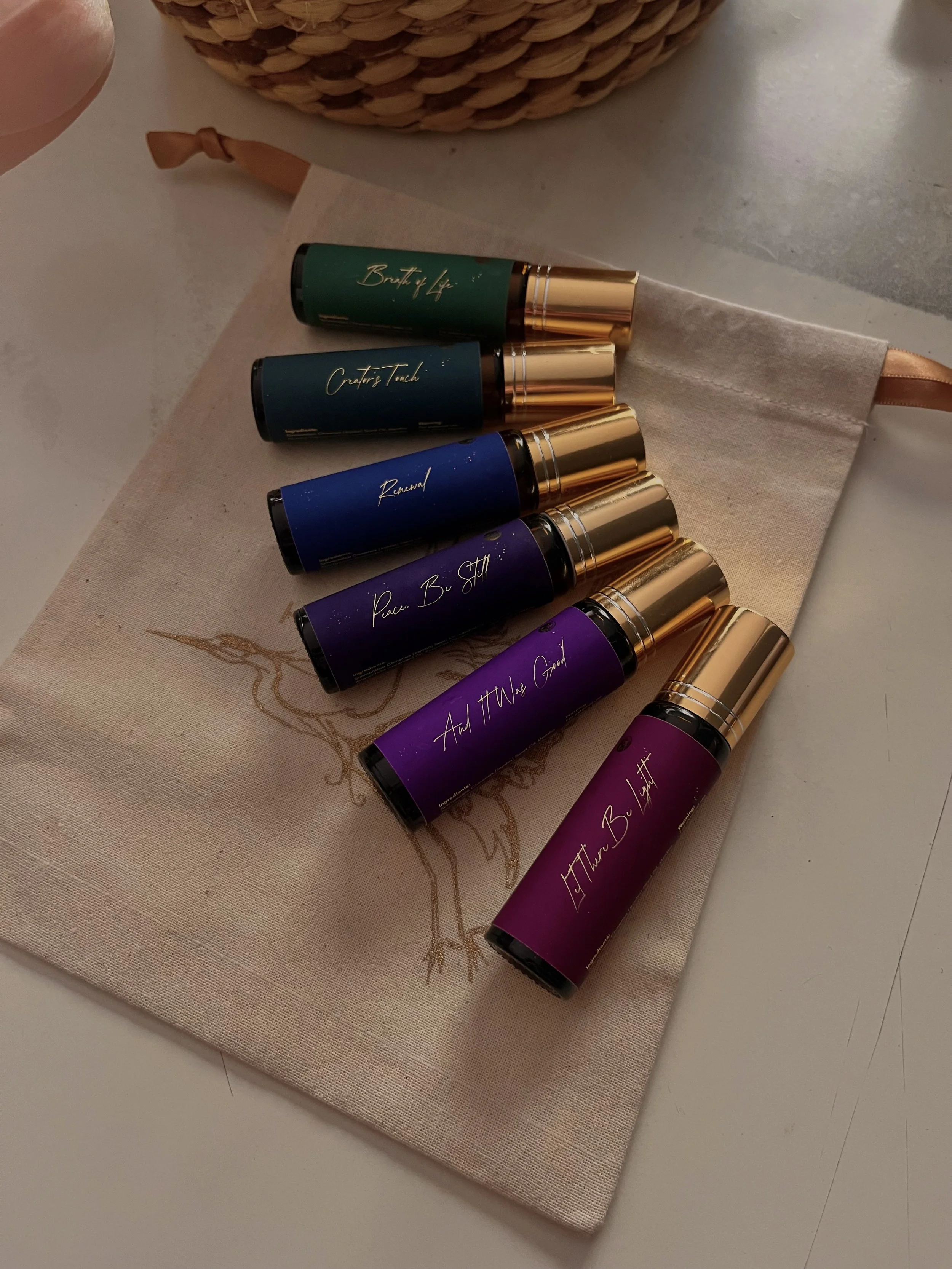 A cloth pouch containing seven colorful essential oils bottles with gold caps, labeled with motivational phrases such as 'Birth of Life', 'Create a Touch', 'Renewal', 'Peace. Be Still', 'And It Was Good', and others, arranged on a beige cloth on a white surface.