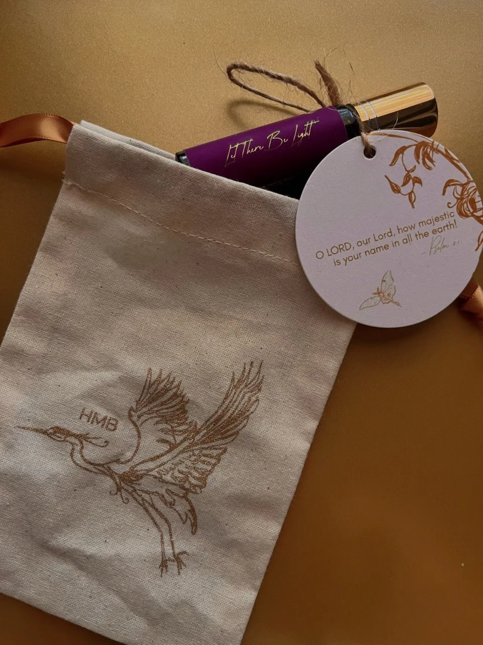 A cream-colored fabric gift bag with a gold illustrated bird design and the initials 'HMB.' Inside the bag is a purple roll-on bottle, a round white tag with gold accents and Christian scripture, and a gold ribbon attached to the bag handle.