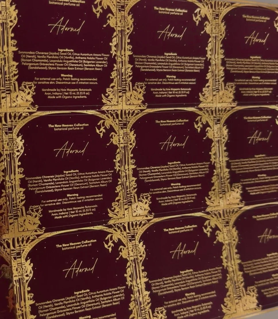 Multiple boxes of botanical perfume oil labeled 'Adorned' from The New Heaven Collection, featuring intricate gold designs and list of natural ingredients.