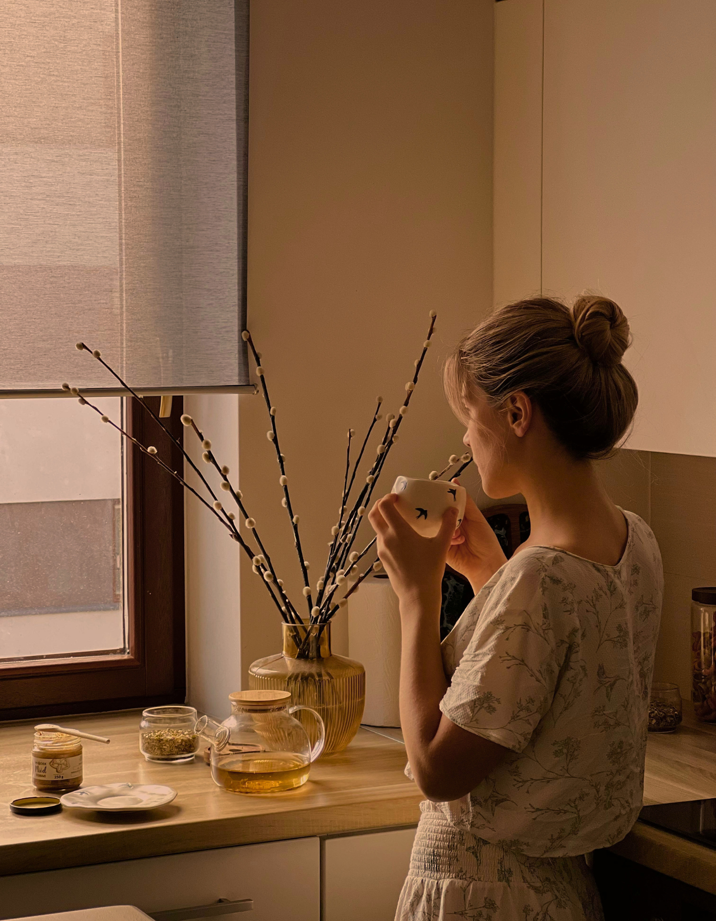 A woman with her hair in a bun is drinking from a mug in a kitchen. There is a window with a roller blind, a yellow vase with pussy willow branches, and various jars and dishes on the kitchen counter. A slow morning.