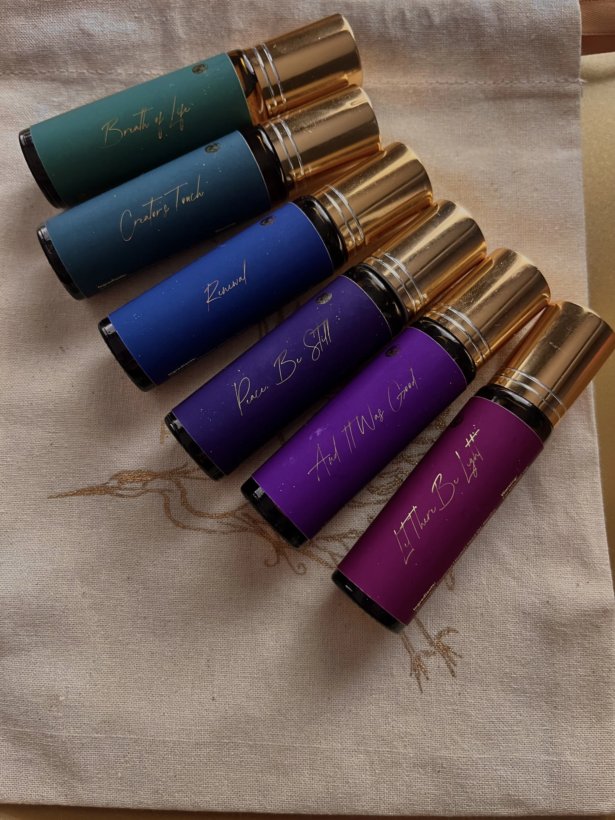 A row of five colored roll-on bottles with gold caps, labeled with different phrases in gold text, resting on a beige fabric surface.