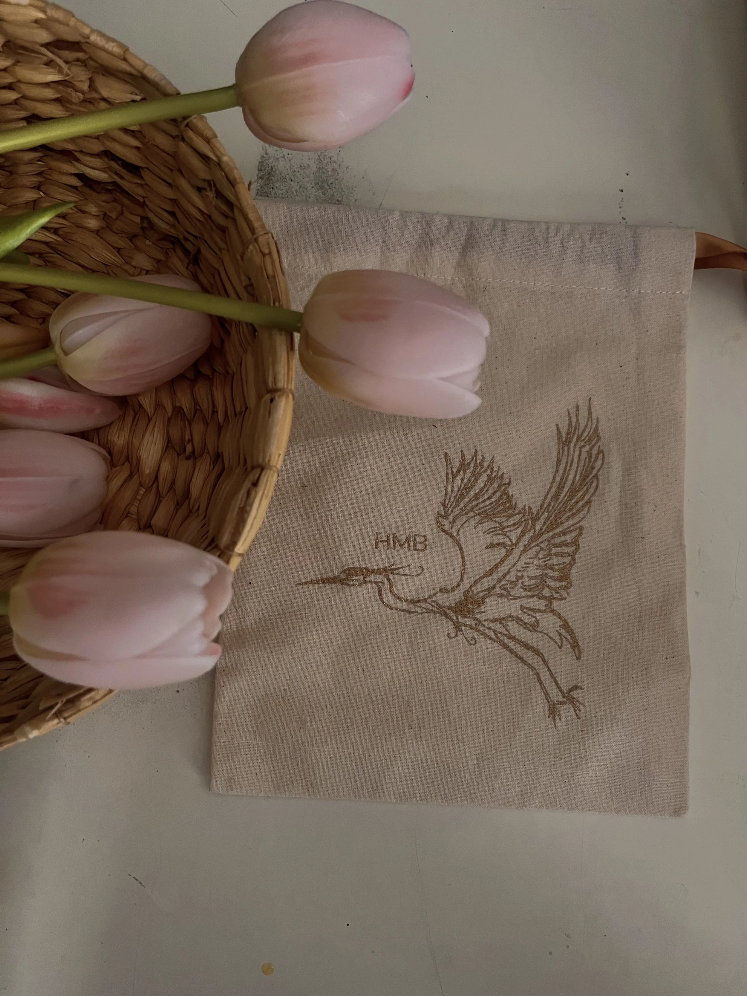 Pink tulips in a woven basket on a table with a beige linen cloth featuring a logo of a stylized bird with the initials 'HMB'.