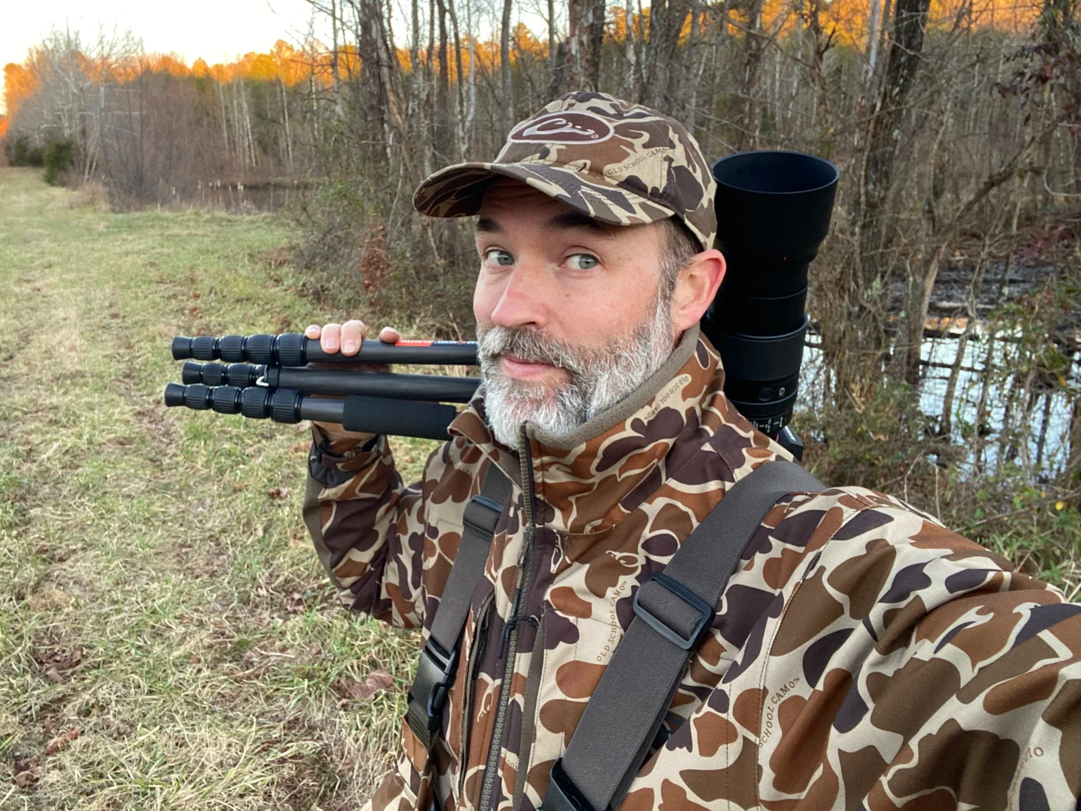 Allen Sherman photography portrait of a man with a gray beard and blue eyes wearing camouflage outdoor clothing and a camouflage hat, standing near a pond at sunset with a tripod on his shoulder and a large camera lens on his backpack, smiling at the