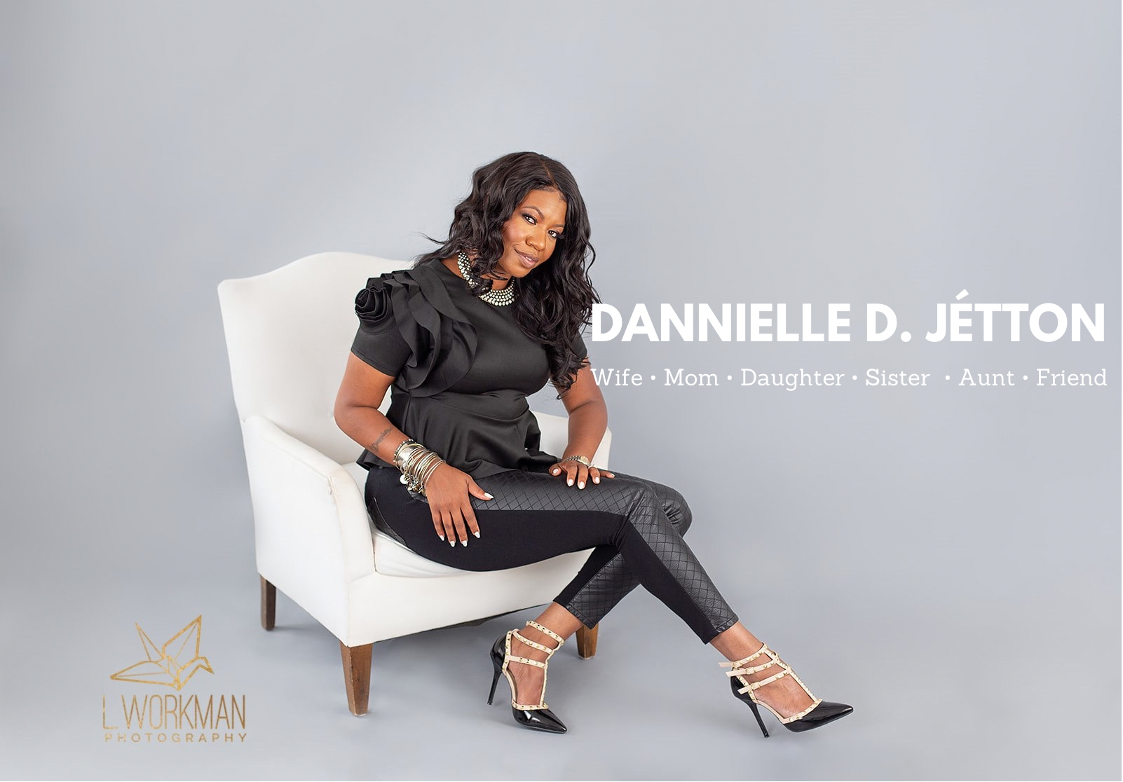 A woman sitting on a white chair against a gray background, wearing a black ruffled top, patterned black pants, and black and beige heels. She has dark hair styled in loose waves, her hands rest on her lap, and she is accessorized with bracelets and rings. Text overlay includes her name, "DANNIELLE D. JÉTTON," and her roles: wife, mom, daughter, sister, aunt, friend. The photograph is credited to L. Workman Photography.