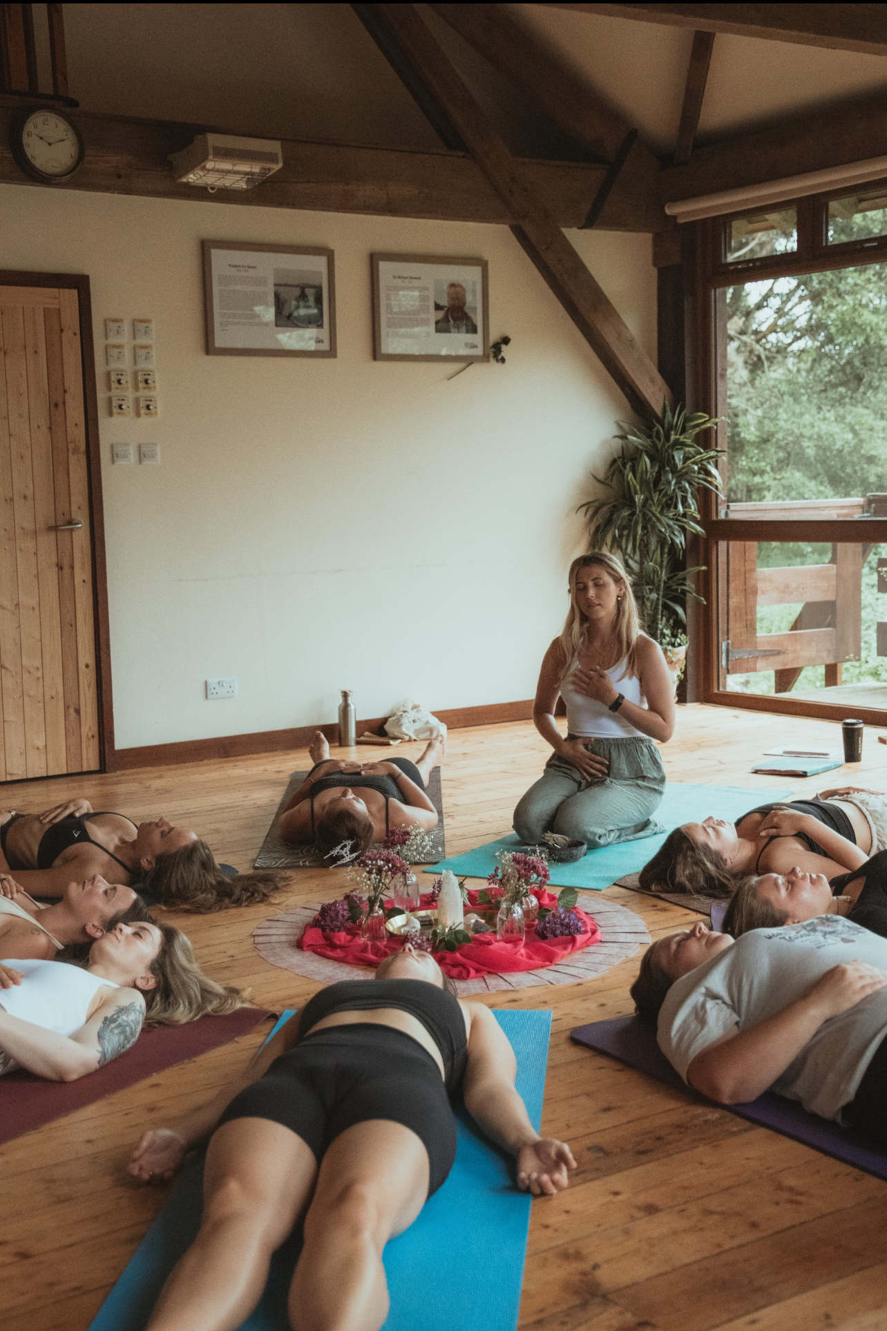 A group of women participating in a yoga or wellness class, lying on yoga mats on a wooden floor in a room with large windows, led by a woman sitting on a mat.