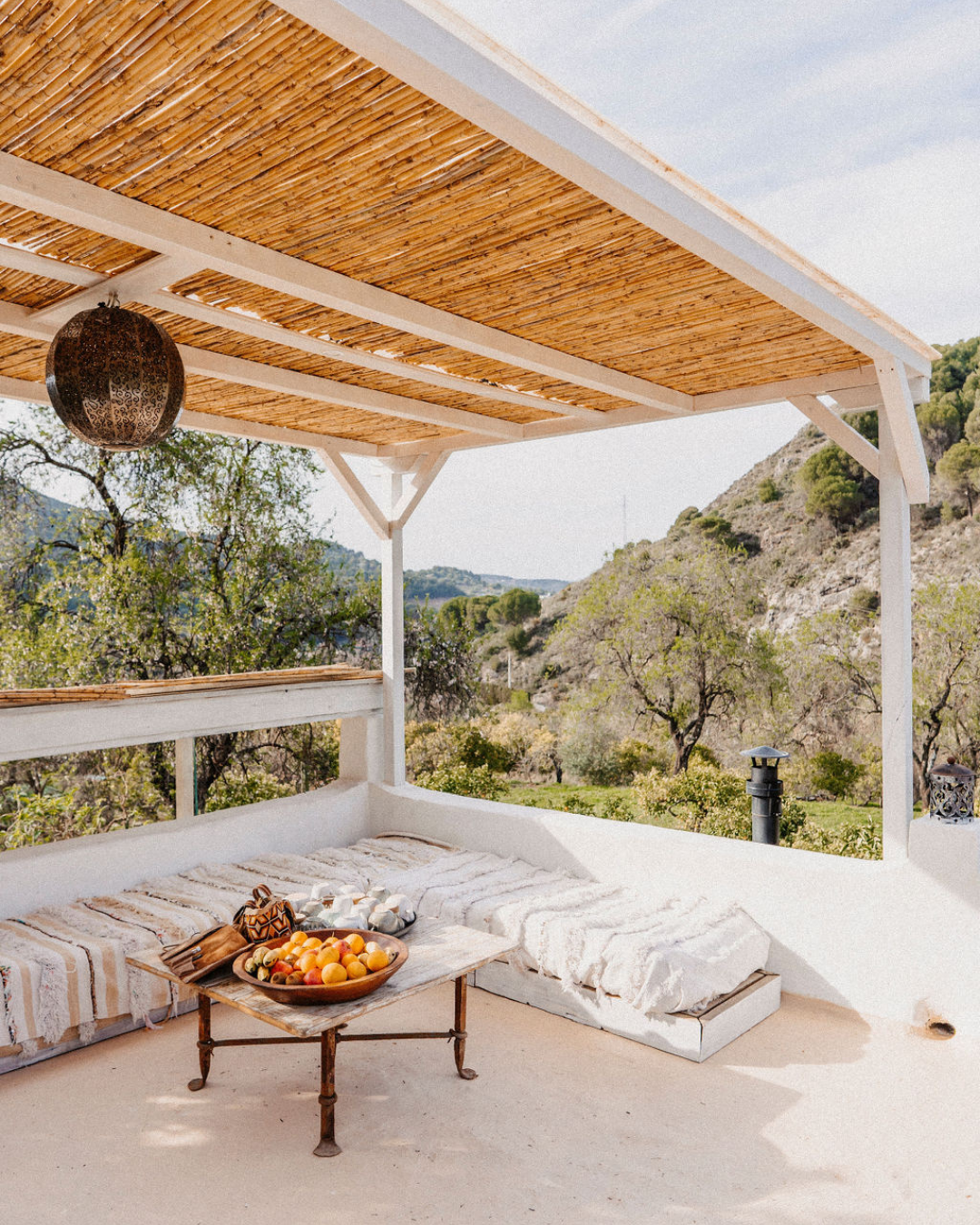 An outdoor patio with a shaded roof, a round wooden table with a bowl of peaches and some other items, a cushioned seating area with a striped fabric, and a scenic background of trees and hills under a partly cloudy sky.