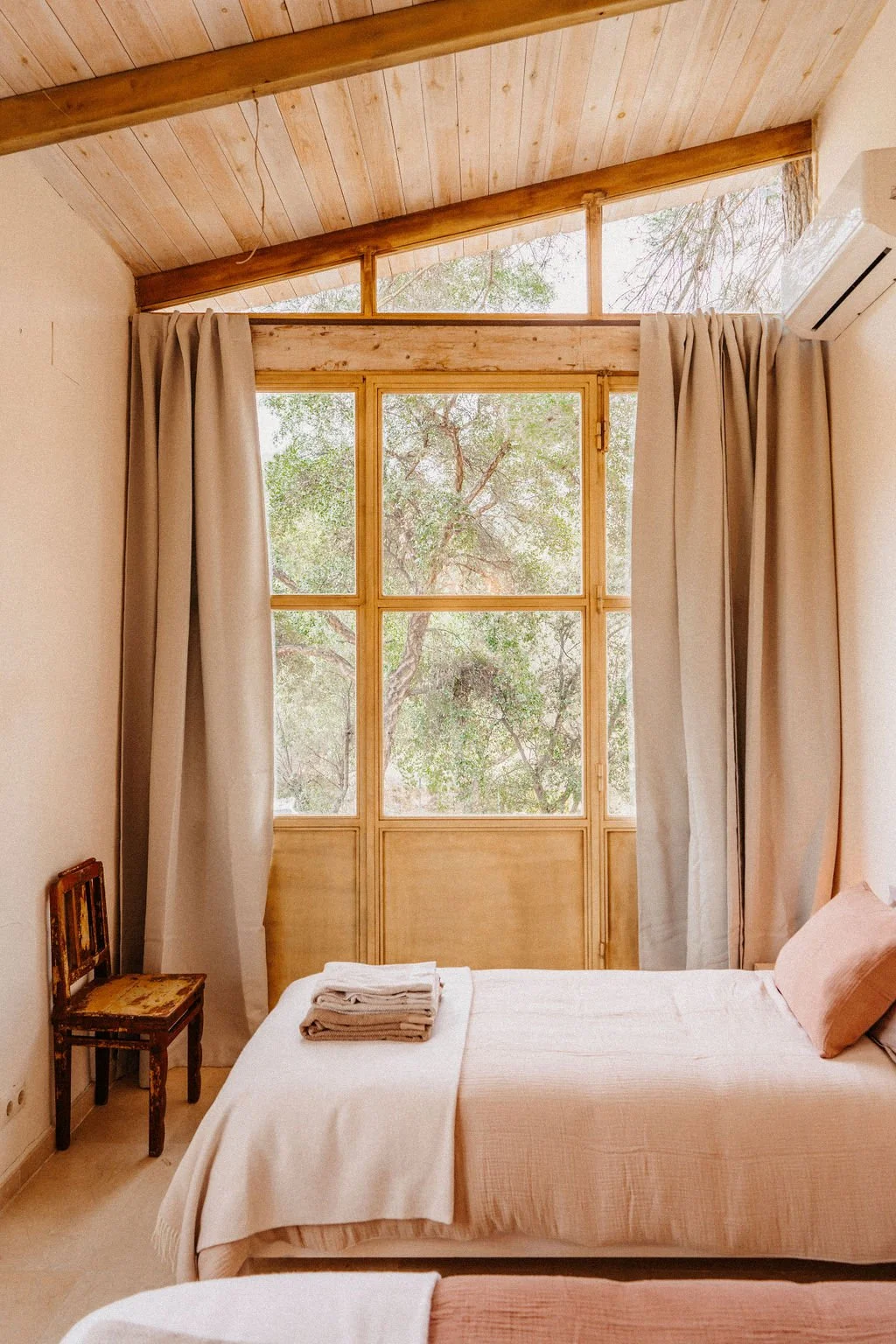 A cozy bedroom with a large window looking out onto trees, beige curtains, a bed with a beige bedspread, and a small vintage chair in the corner.