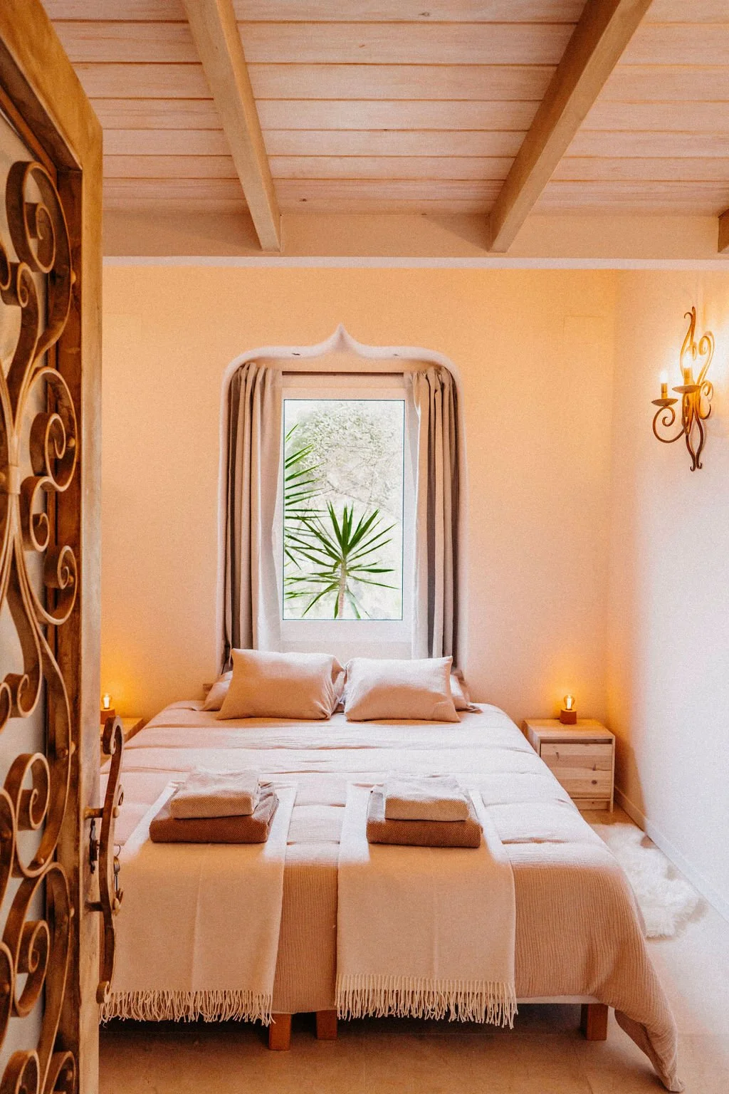 A cozy bedroom with a large bed, two pillows, and folded towels at the foot. There is a window with curtains and a green plant outside. Soft lighting from small lamps on nightstands and a wall sconce create warm ambiance.