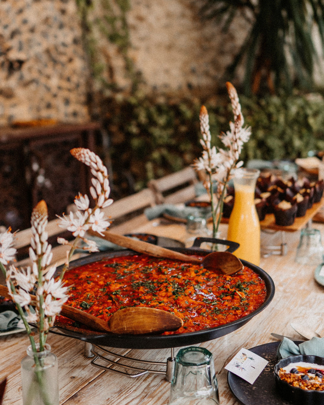A table set with a large skillet of tomato-based stew or sauce, flowers in vases, a bottle of juice, and smaller dishes in the background, with a rustic and cozy ambiance.