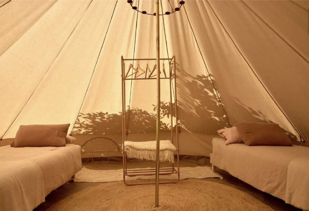 Interior of a beige canvas tent with two couches on either side, pillows, a rug on the floor, and a central clothing rack with a blanket on it, casting shadows of shadowy foliage on the tent walls.