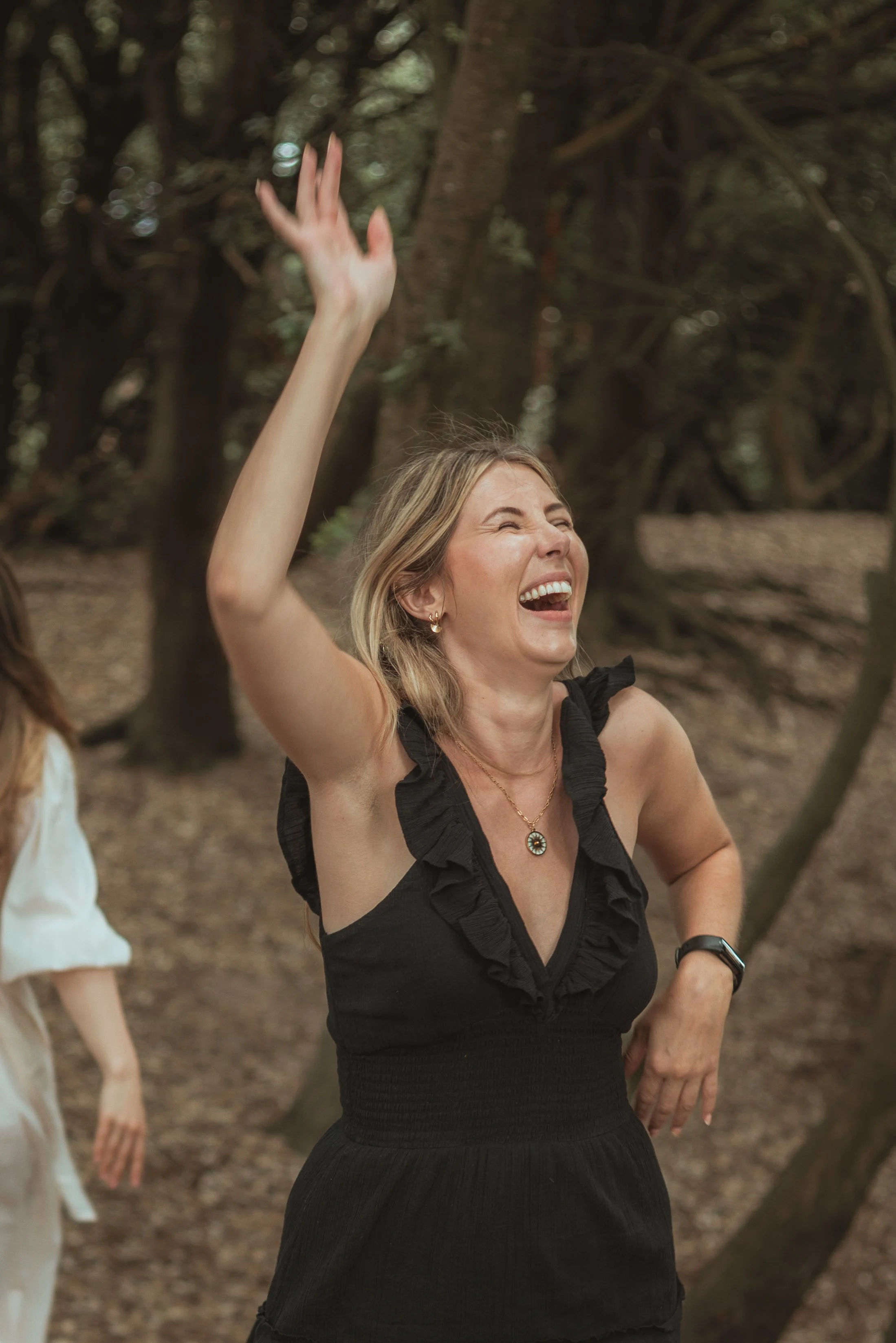 Woman in black dress laughing and raising her hand in an outdoor wooded area.