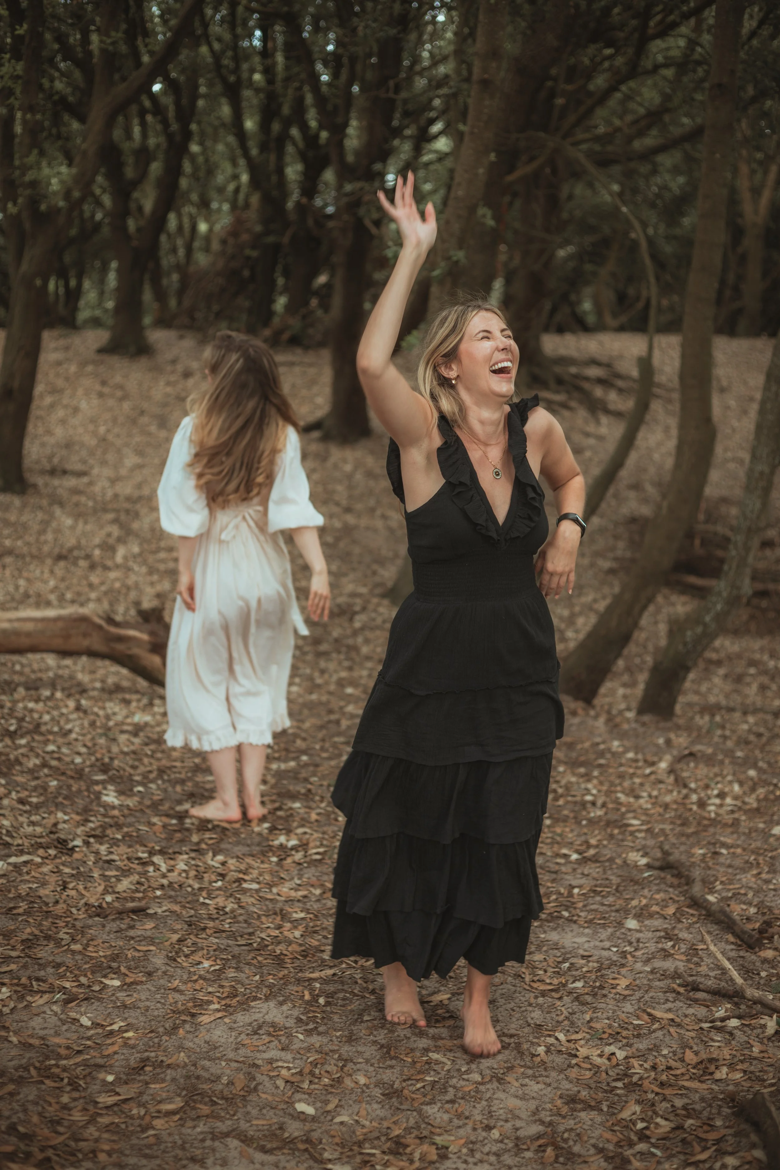 A woman in a black dress laughing and raising her hand while walking barefoot on a wooded trail, celebrating at a women's circle