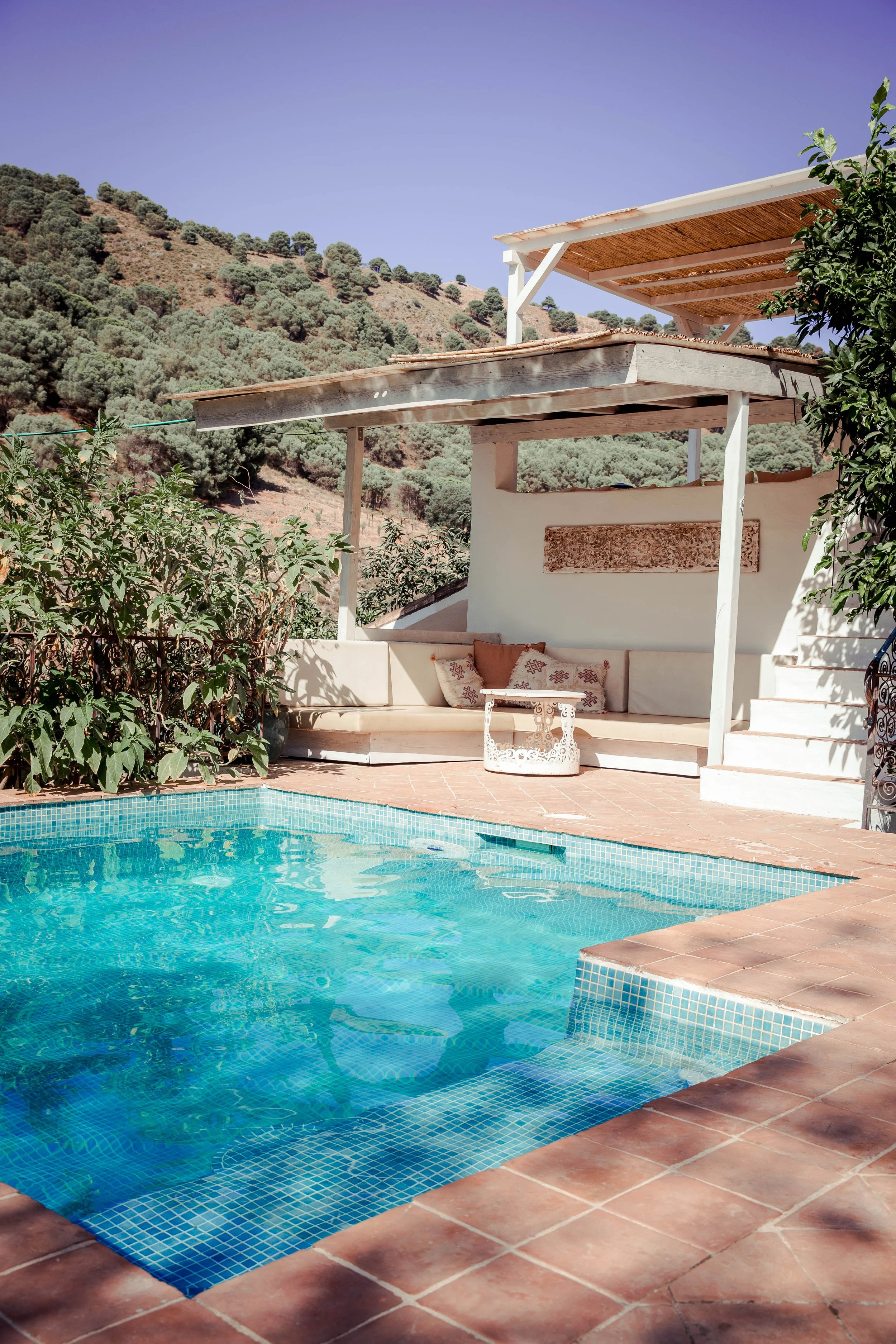 A small outdoor swimming pool with blue tiles, surrounded by terracotta tiles, overlooking a hillside with green trees, and featuring a shaded seating area with cushions and a small table.
