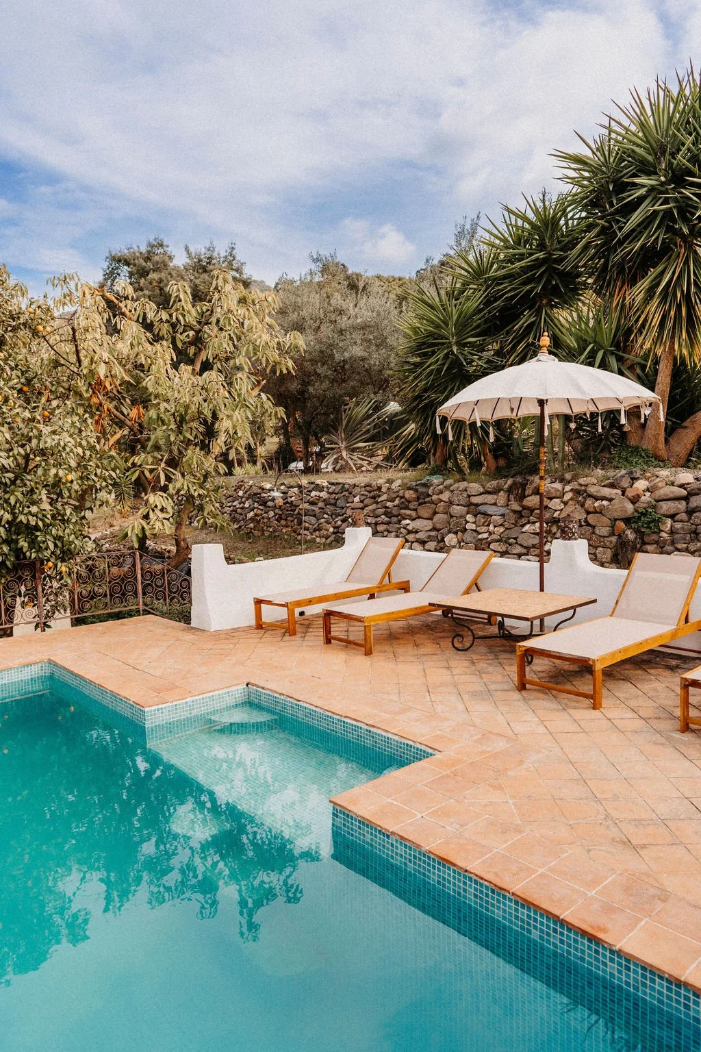 A backyard with a swimming pool, lounge chairs, a white umbrella, and lush green trees under a partly cloudy sky.