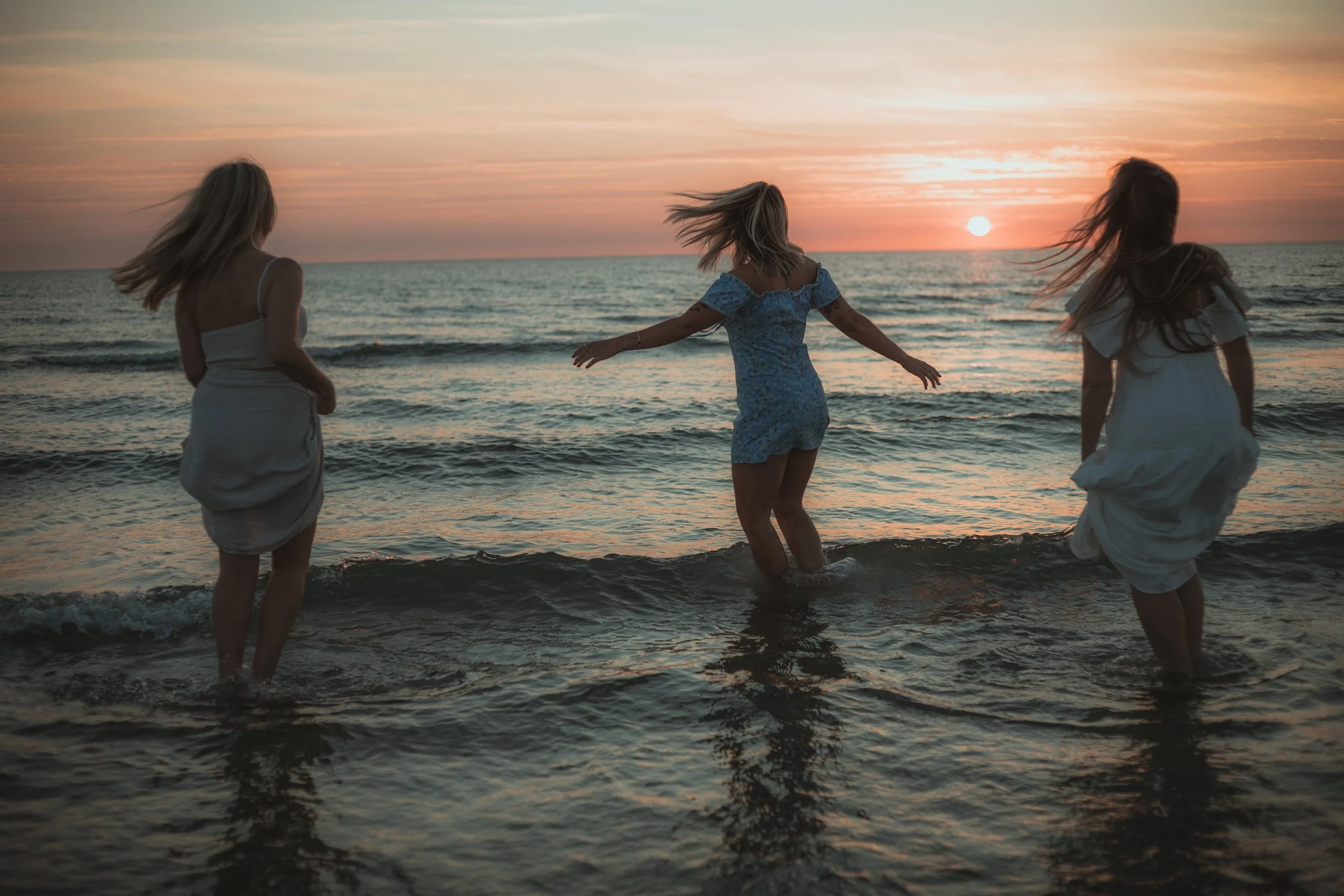 Three women in dresses enjoying themselves in the shallow ocean water during sunset at the beach, with their hair flowing in the breeze.