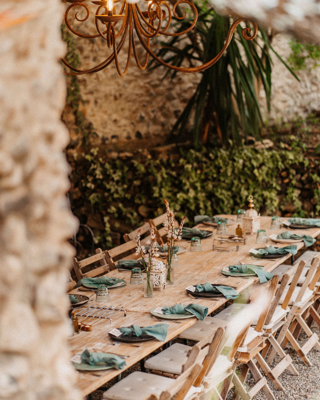 Long wooden table set for a gathering with napkins, glasses, and decorative centerpieces, outdoors under a chandelier.
