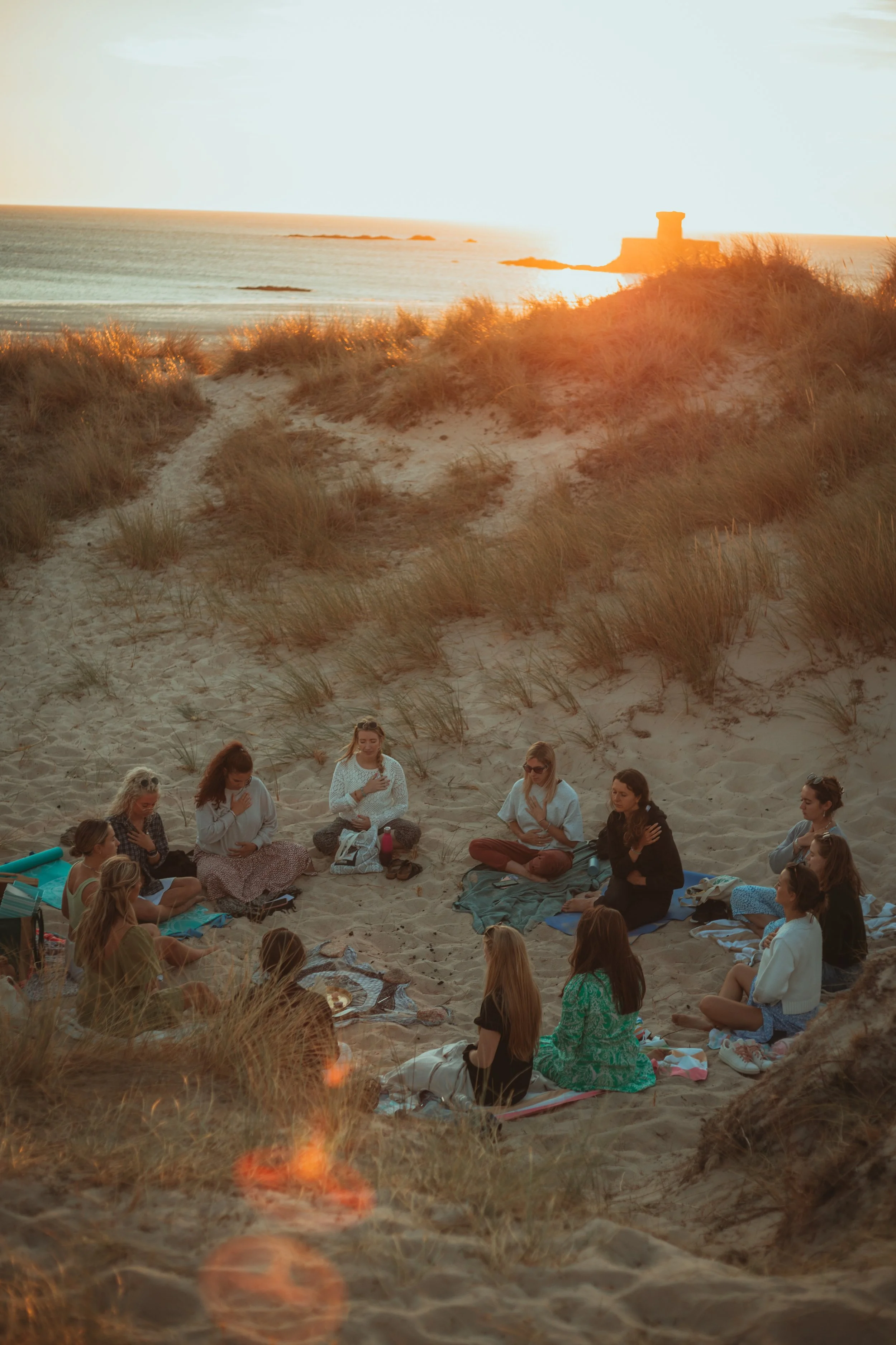 A group of people participating in a meditation or prayer session on a sandy beach at sunset, sitting on blankets and facing each other, with dunes and a distant tower in the background.