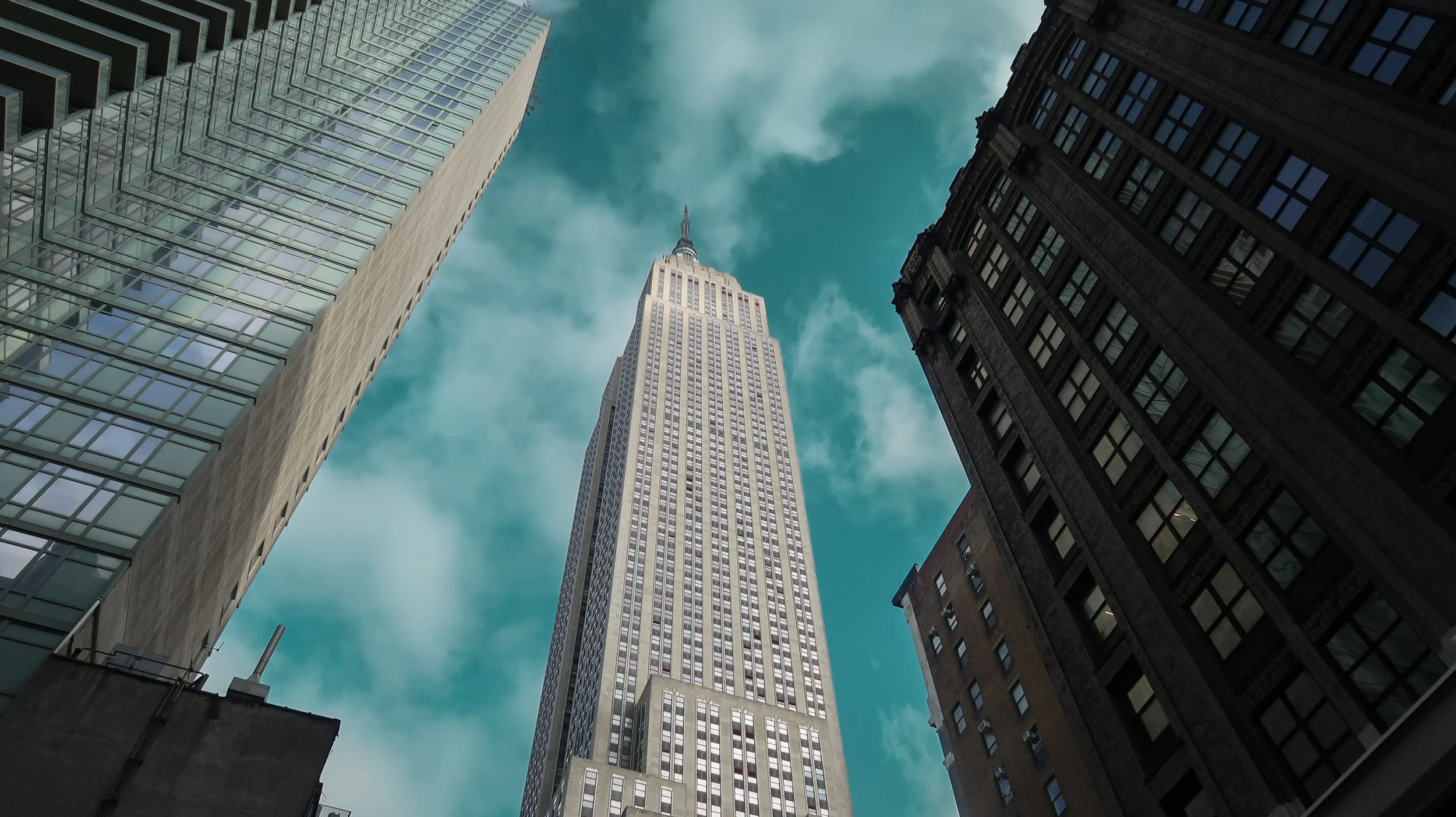 A view looking up at the Empire State Building flanked by tall glass and brick skyscrapers against a partly cloudy sky.