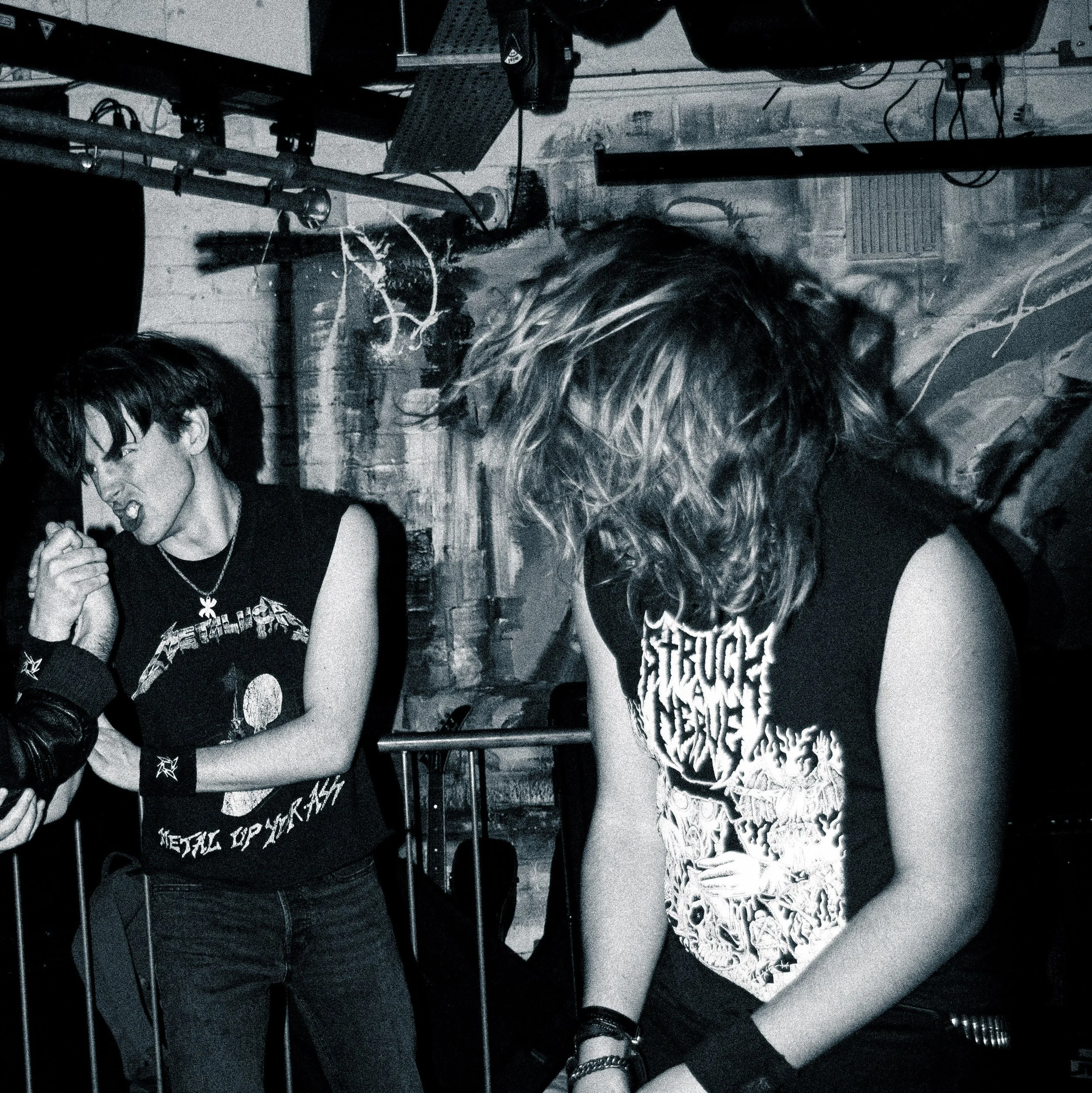 Two young men with long hair performing at a music venue, wearing metal band shirts, with exposed brick walls and music equipment in the background.