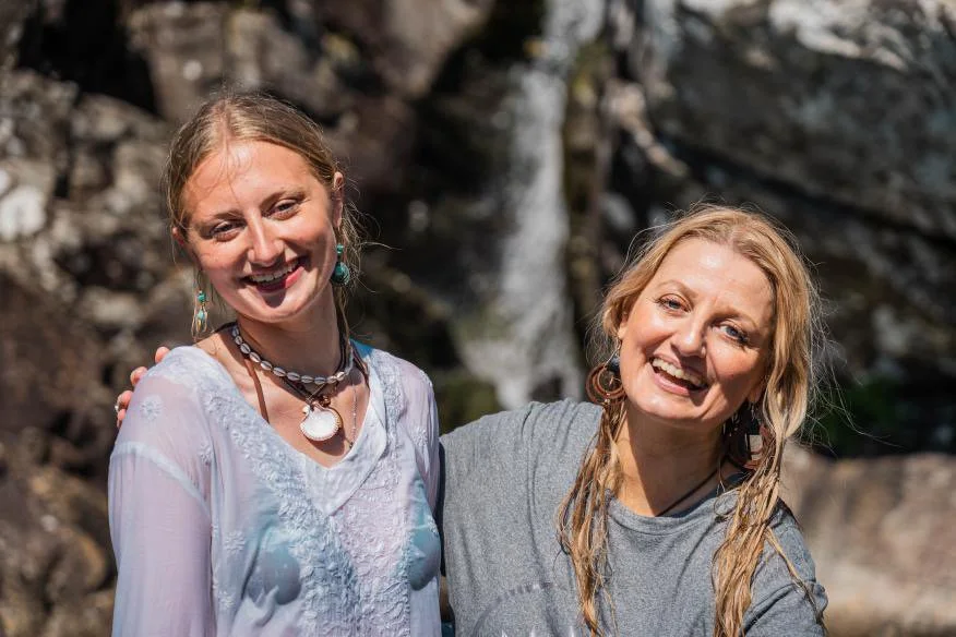 Two women smiling and standing close together outdoors with rocky background and water stream.