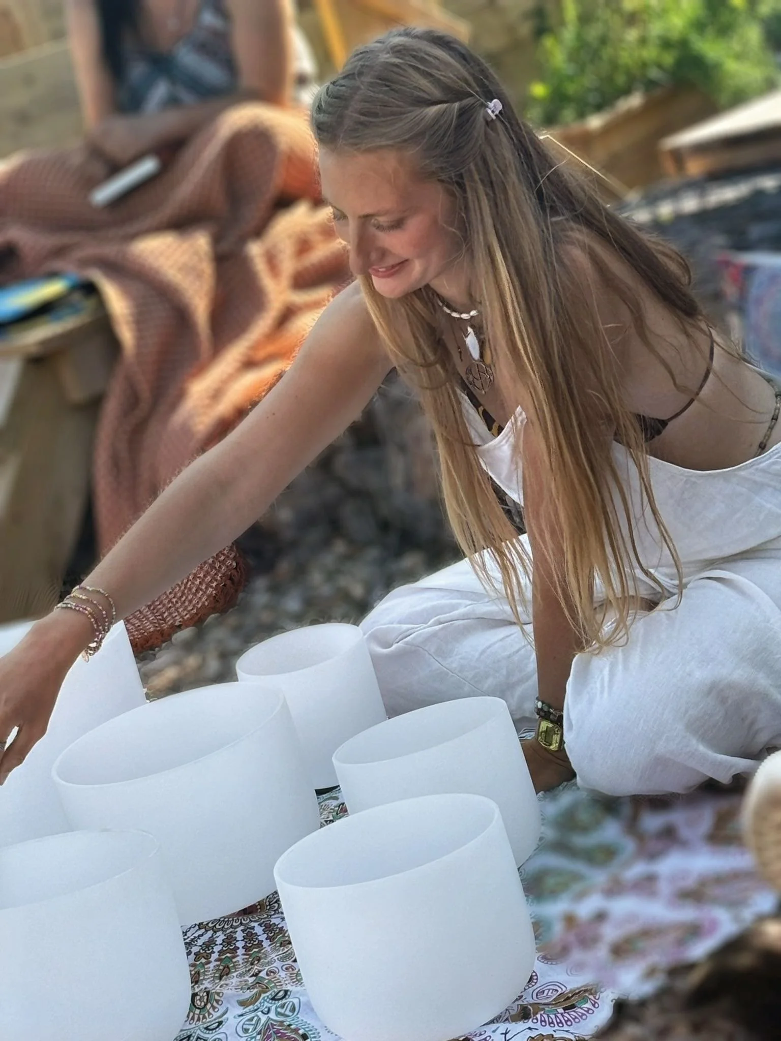A young woman with long blonde hair kneeling outdoors, playing crystal singing bowls on a patterned cloth. She is smiling and wearing a white outfit with jewelry.