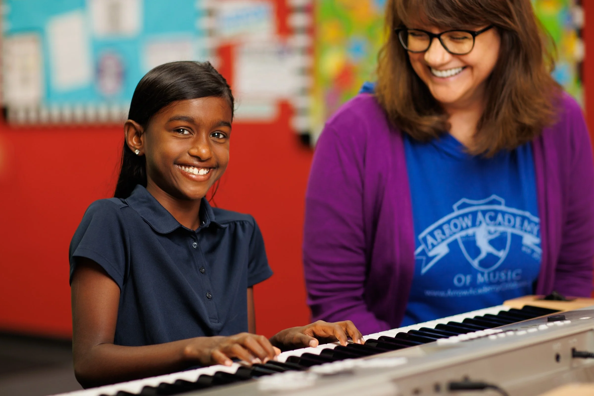 A young girl smiling while playing a keyboard, with a woman beside her, also smiling in a music classroom.