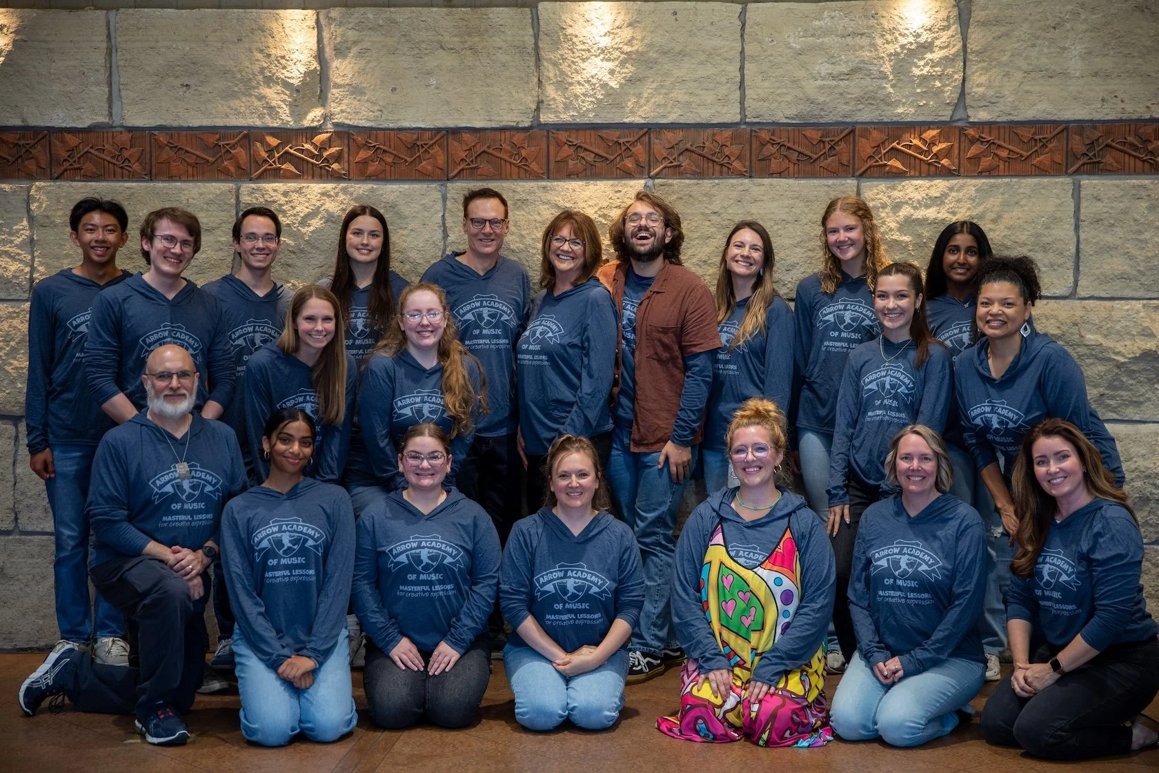A group photo of 20 people, including students and teachers, posing together indoors against a stone wall. Most individuals are wearing matching blue hoodies with a logo that says 'Arrow Academy of Music.' One person in the front row is wearing a colorful dress with bright patterns.