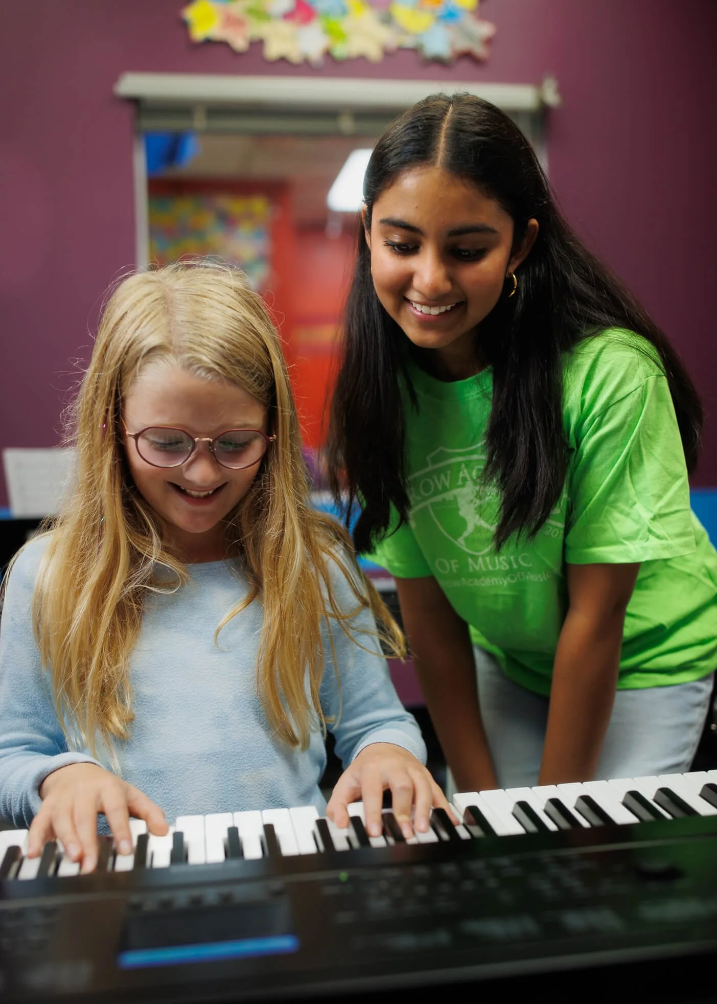 Arrow student assistant helping a younger student at the piano with piano classroom in the background