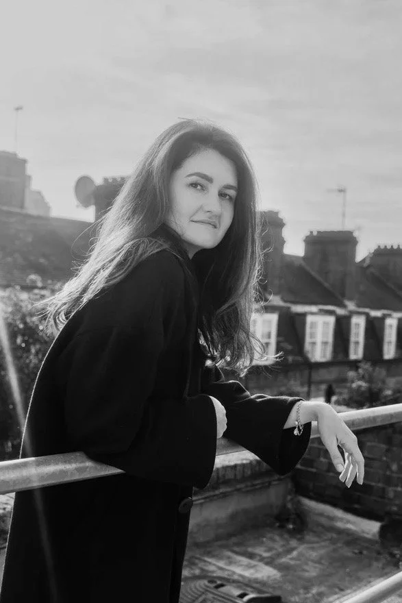 A young woman with long hair standing on a rooftop balcony, looking at the camera, wearing a dark coat in black and white.