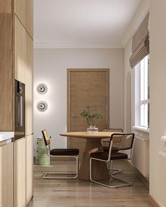 Dining area with wooden round table, three chairs with black cushions, a small plant in a glass vase, and two wall-mounted lights on a white wall near a wooden door, adjacent to a window with a brown Roman shade.
