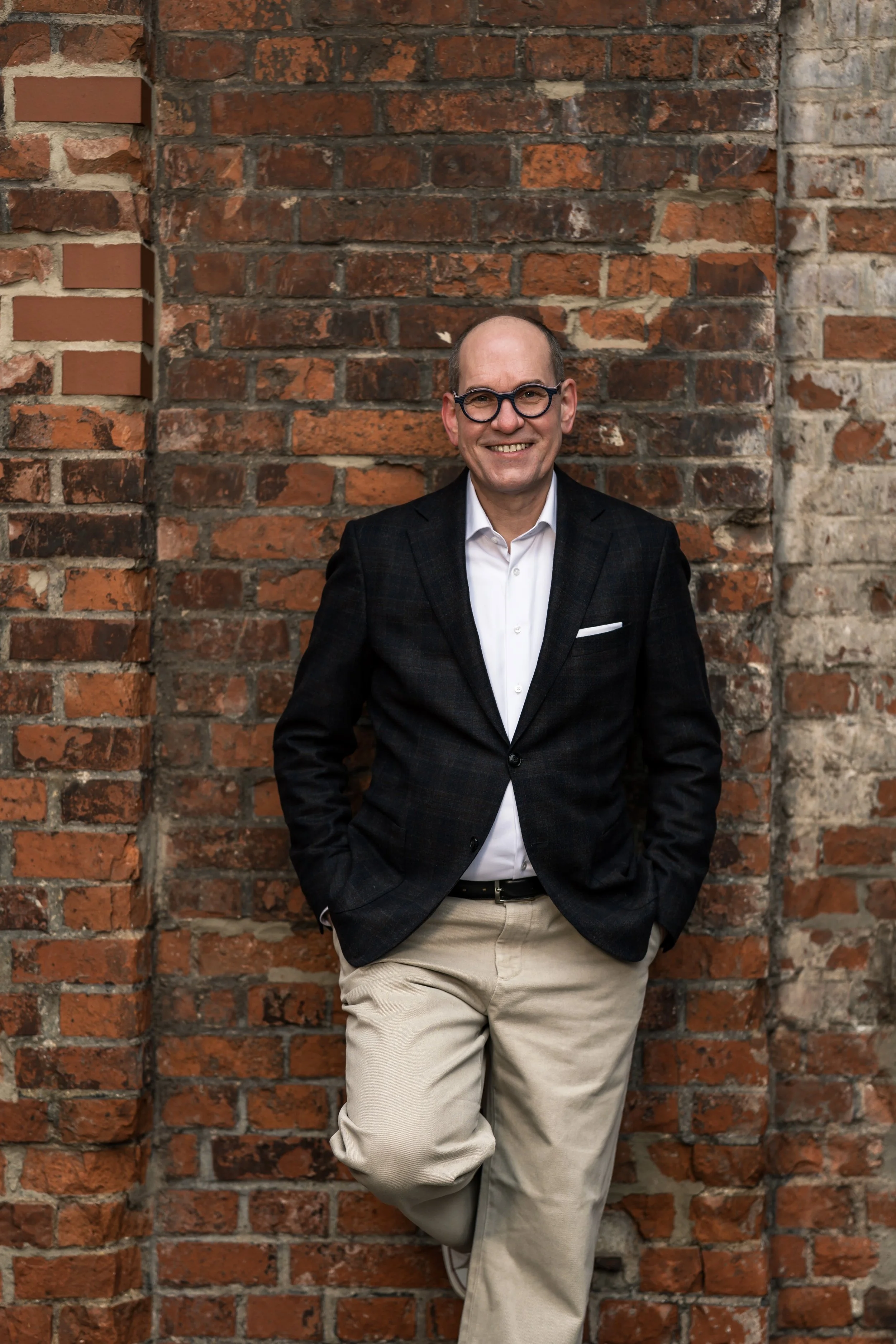 Ein Mann trägt Jacket, gepflegt, leger, weißes Hemd und beige Hosen, steht vor einer alten Backsteinmauer, lächelt und hat die Hände in den Taschen, amerikanische Pose.