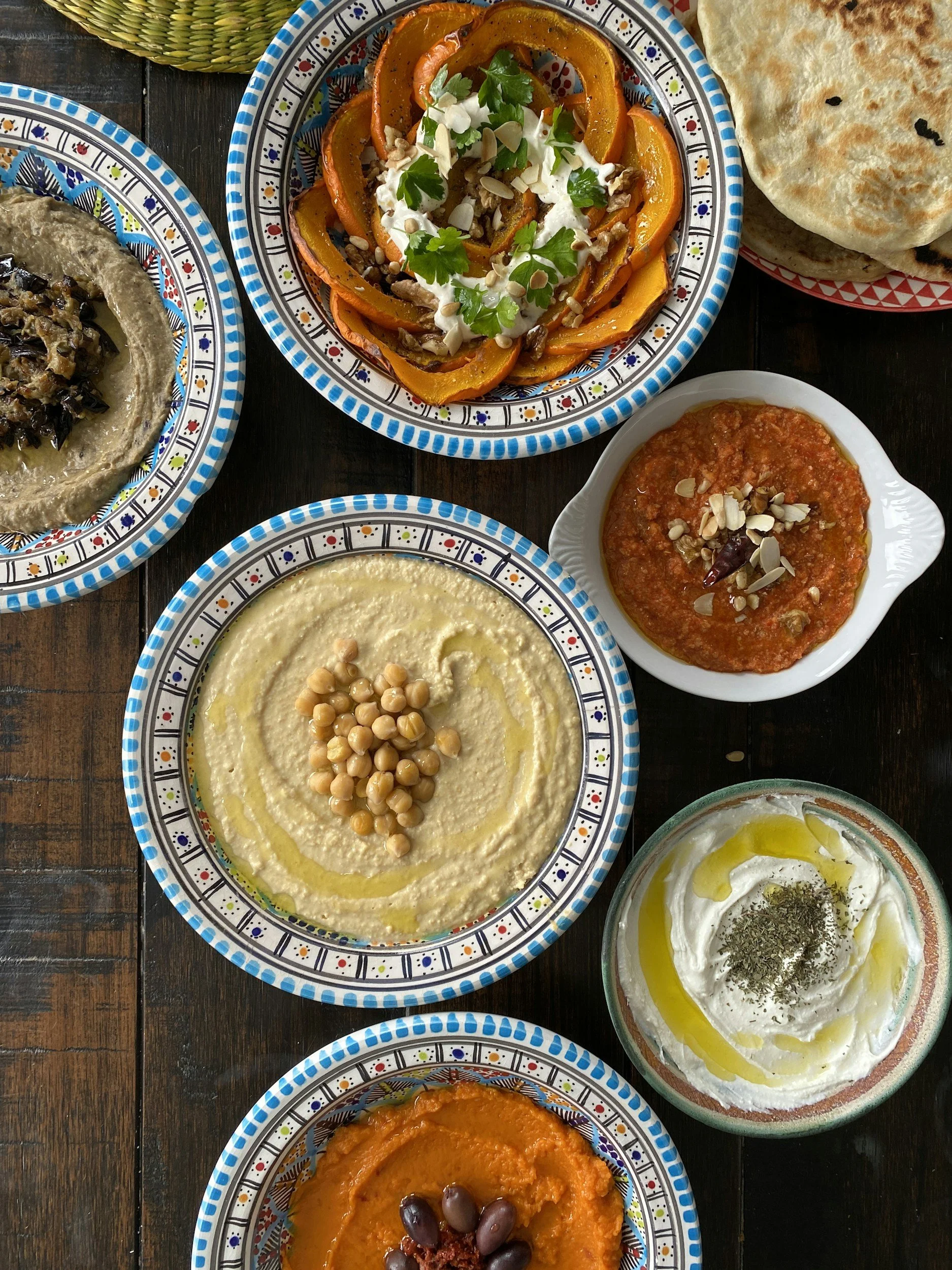 Verschiedene traditionelle arabische Gerichte auf bunten Tellern, darunter Hummus, Moutabal, Baba Ghanoush, aush, und Quark mit Olivenöl und Kräutern, serviert auf einem Holztablett.