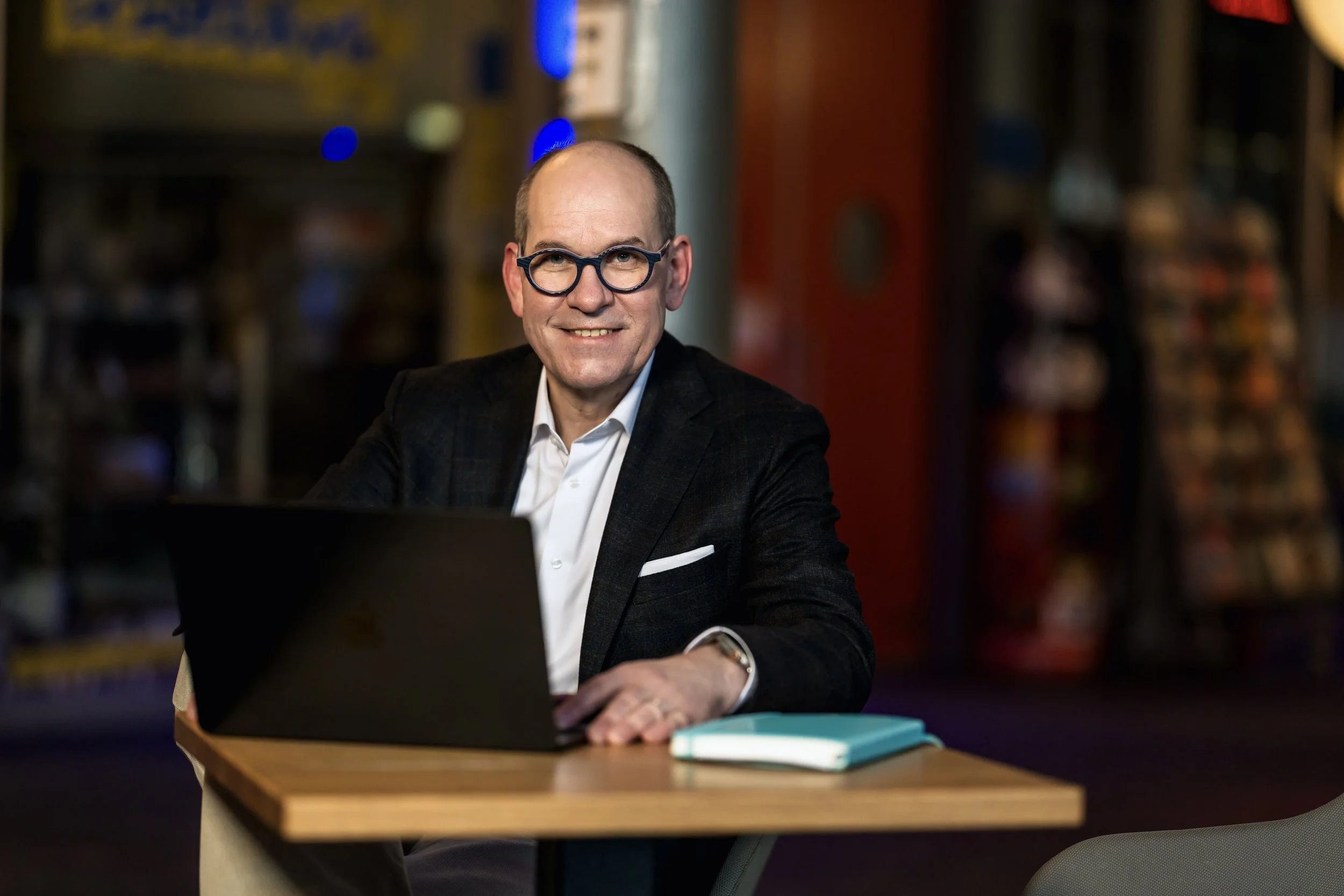 Ein Mann mit Jacket sitzt an einem Tisch in einem Restaurant oder Café, arbeitet an einem Laptop, trägt eine Brille und hat einen aufgeräumten Arbeitsplatz mit einem Notizbuch.