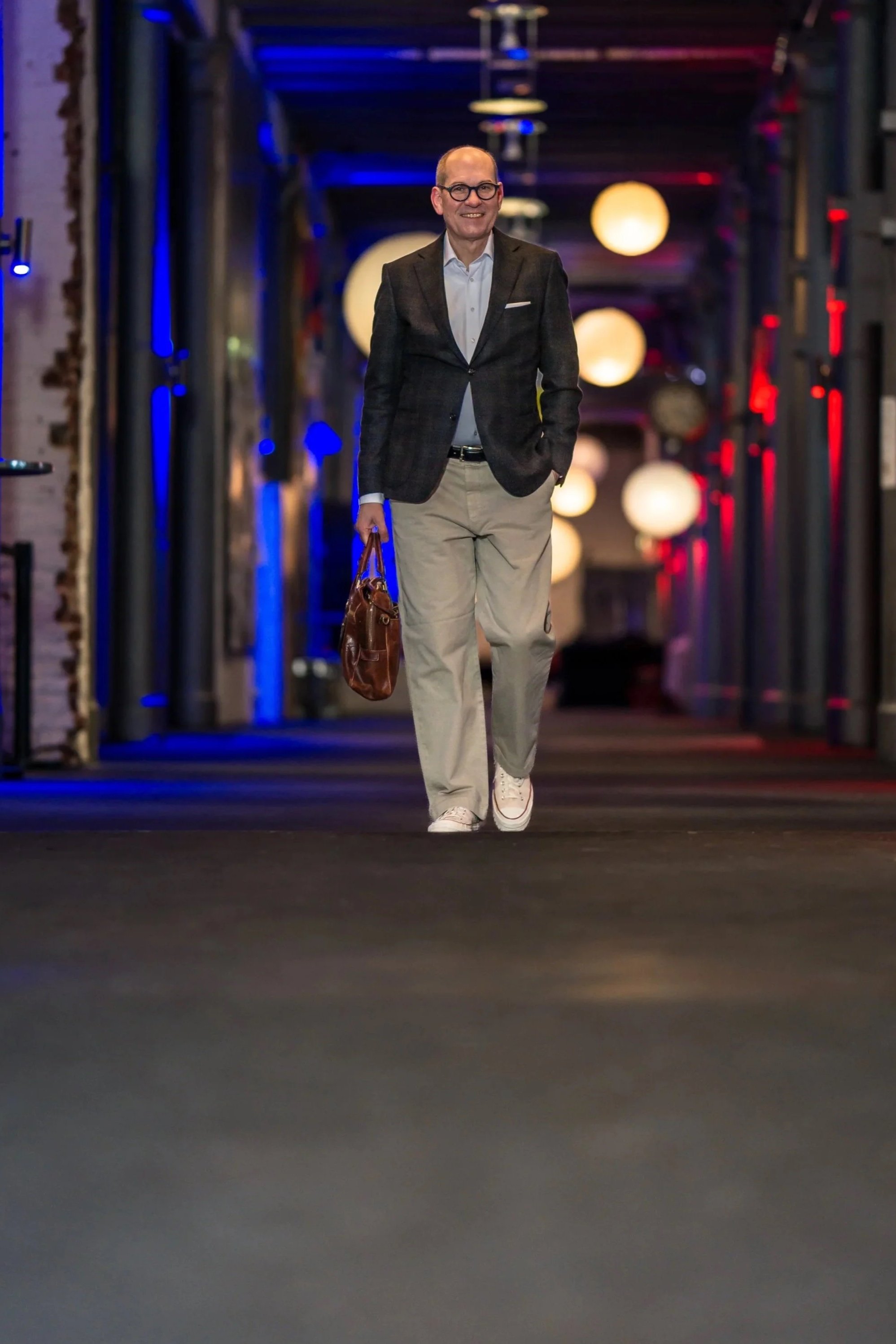 Ein Mann in Business-Kleidung, der in einem gut beleuchteten, modernen Flur oder Korridor spaziert, lächelnd, mit Brille, dunklem Jacket, weißen Hemd, beigefarbenen Hosen und weißen Chucks, trägt eine braune Tasche.