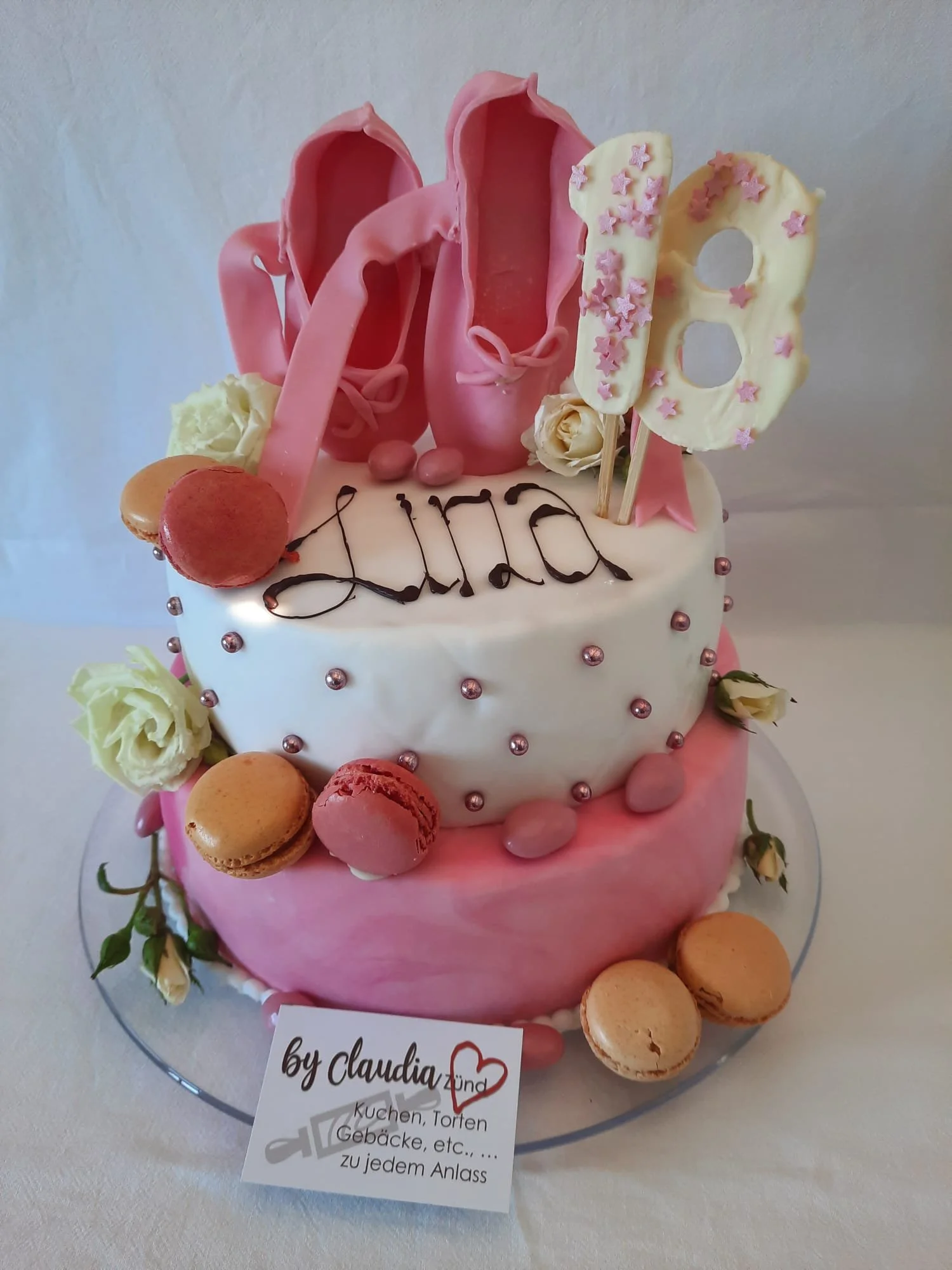 Zwei-tiers Geburtstagstorte mit rosa und weißer Dekoration, Macarons, weißen Rosen und einem Bewerbungskuchen mit dem Namen 'Lina' sowie der Zahl '18' als Dekoration.