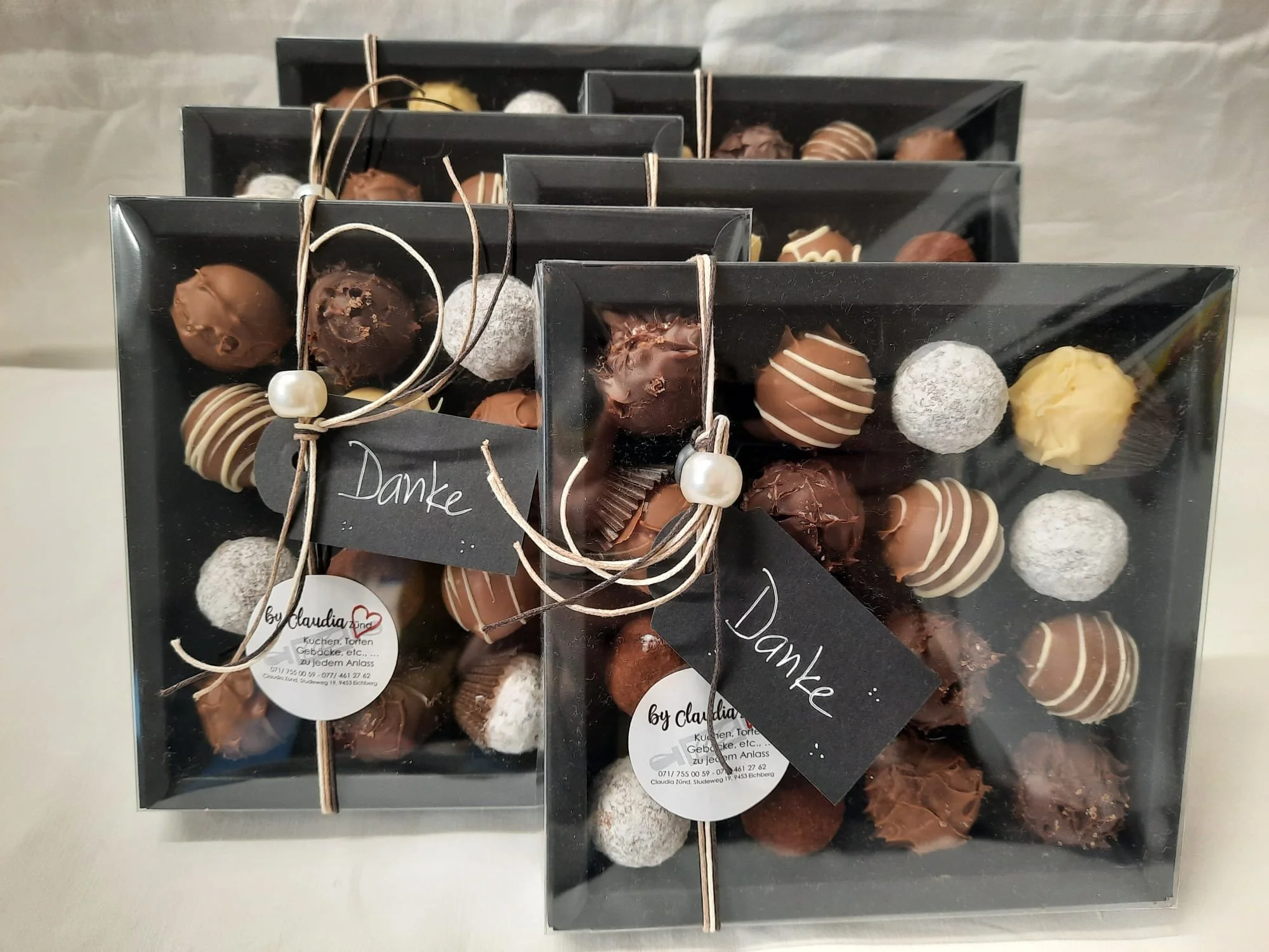 Geschenkboxen mit assortierten Pralinen, verziert mit kleinen schwarzen Schleifen und einer Dankeskarte, auf silbernem Hintergrund.