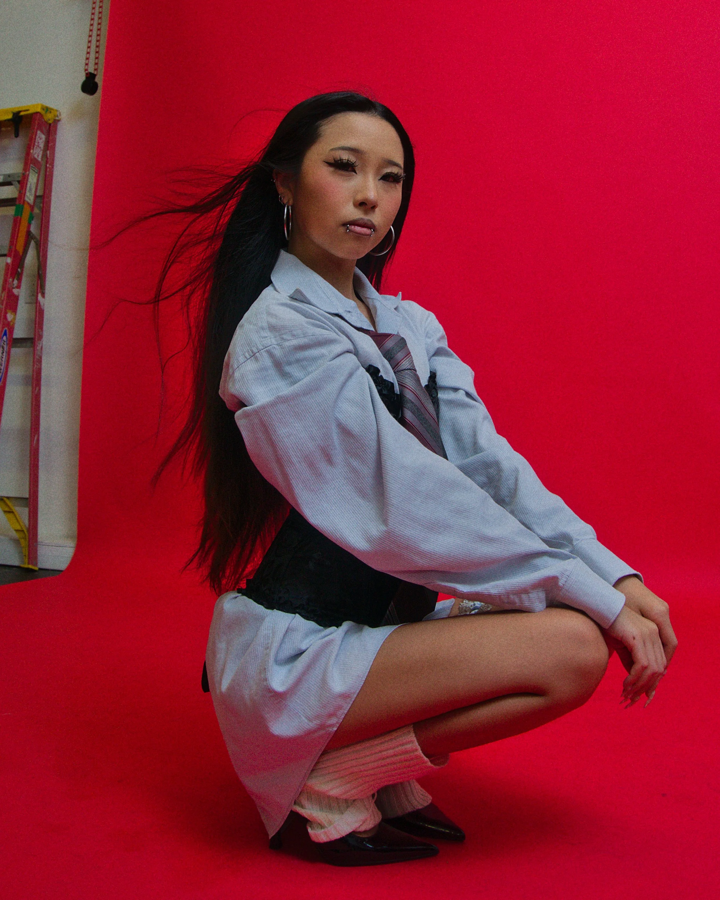 A young woman with long black hair, wearing a school uniform with a long sleeve shirt, plaid tie, and black waist corset, squatting against a bright red backdrop in a studio setting.  Photographed by Erin De Shay.
