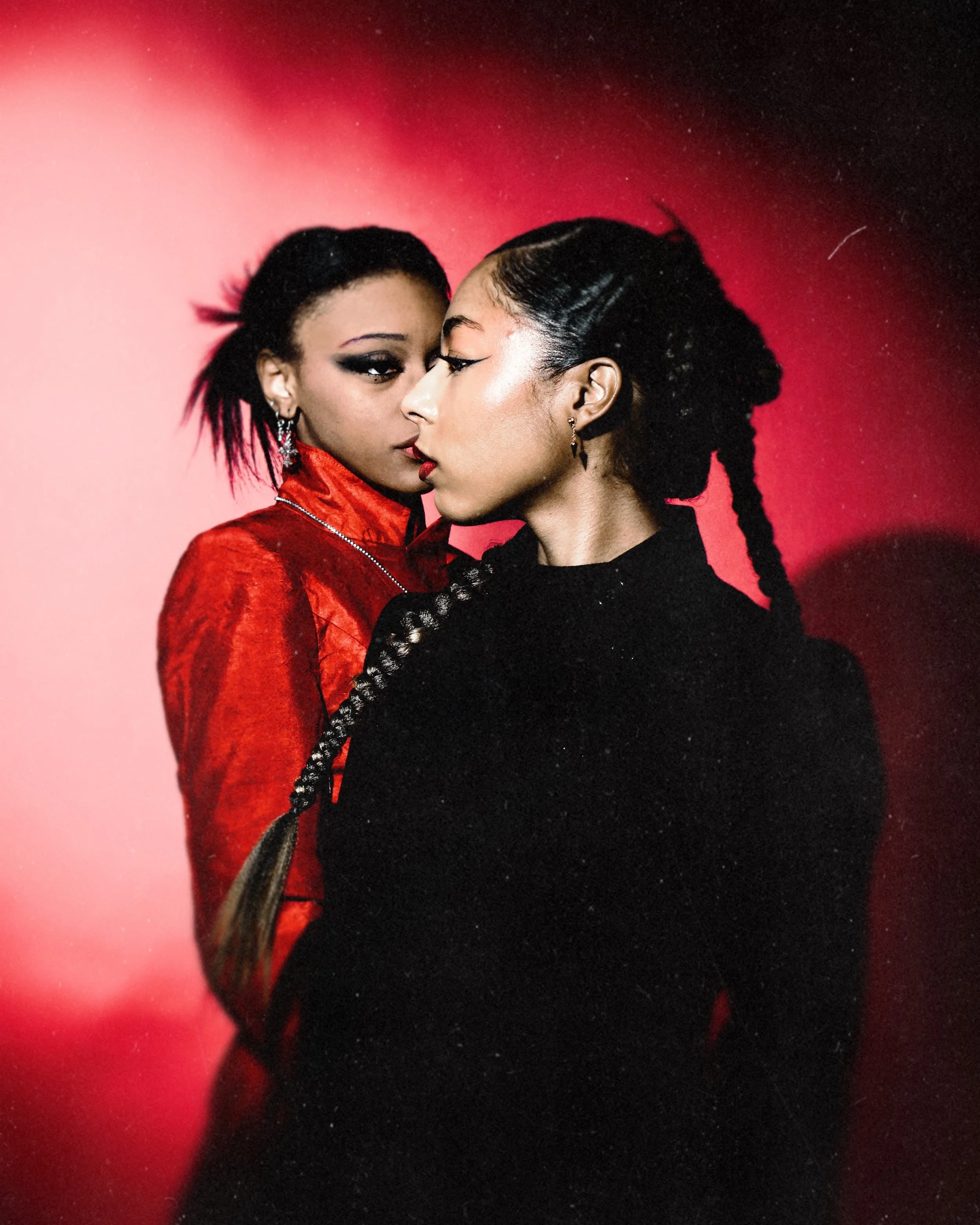 Two women with braided hair, facing each other closely against a red-pink gradient background.  Photographed by Erin De Shay.